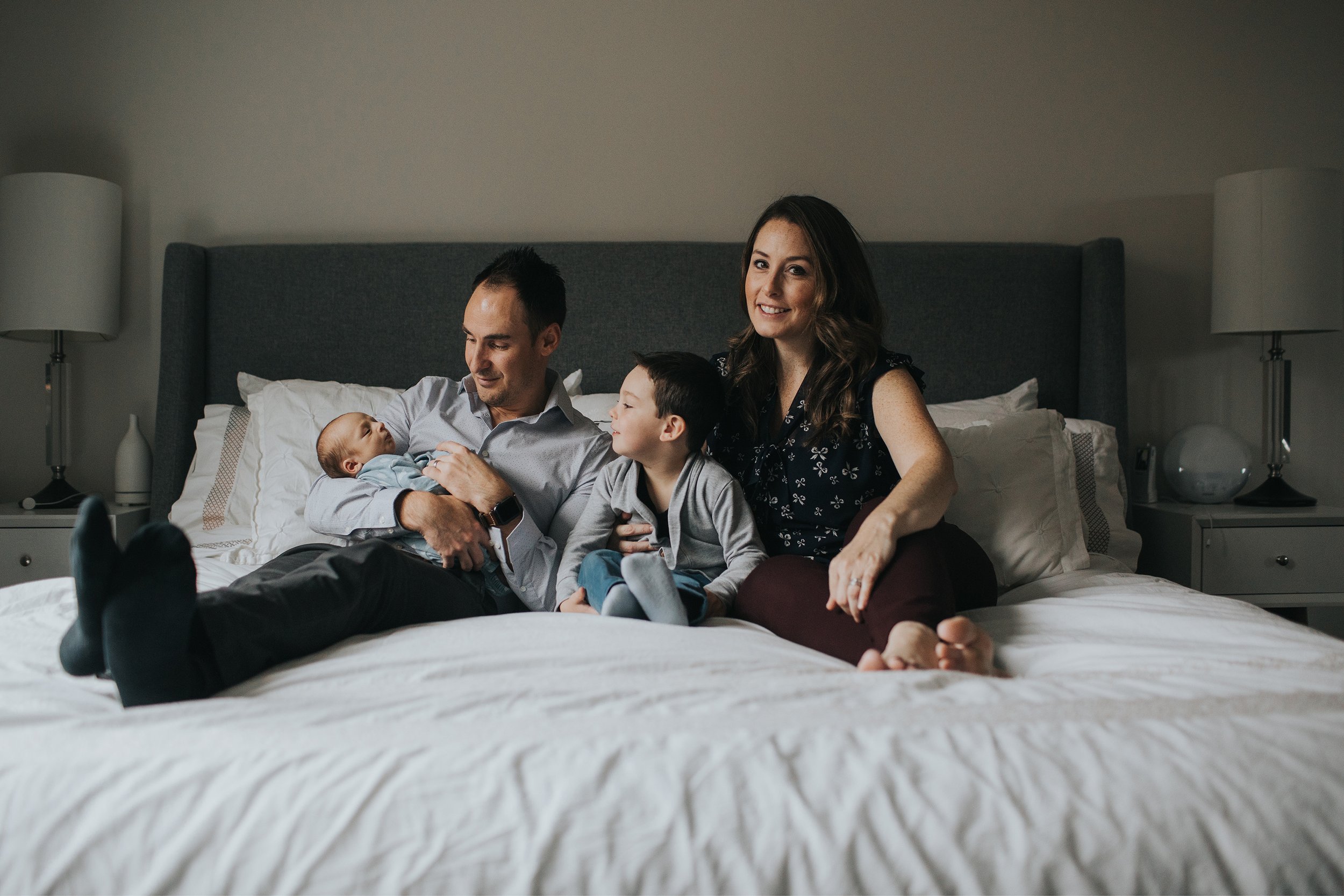 A candid family moment with parents and their children, including a newborn, sitting together on a bed.