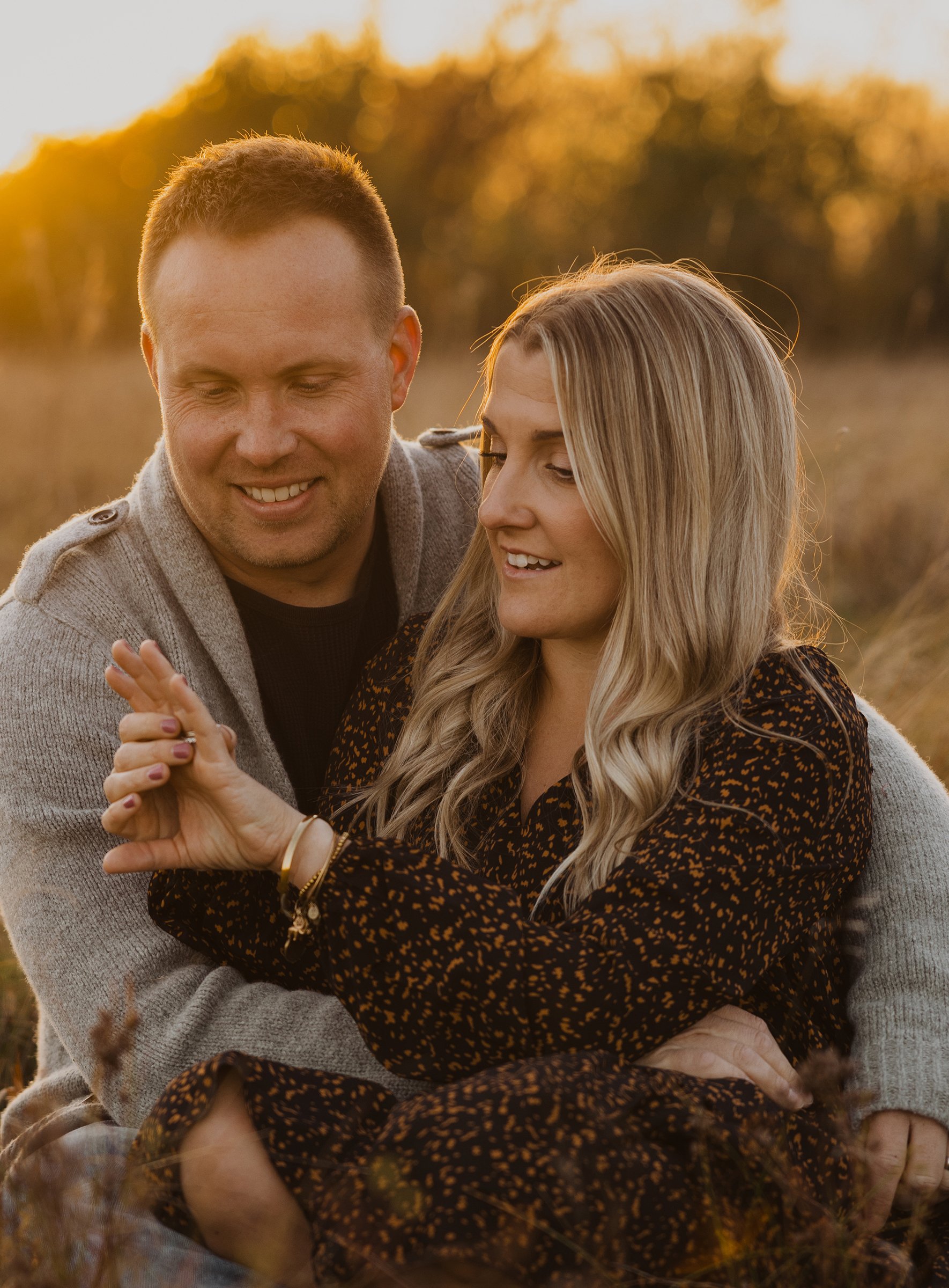 A couple embraces in a warm sunset glow while seated in a field, capturing a tender, romantic moment.