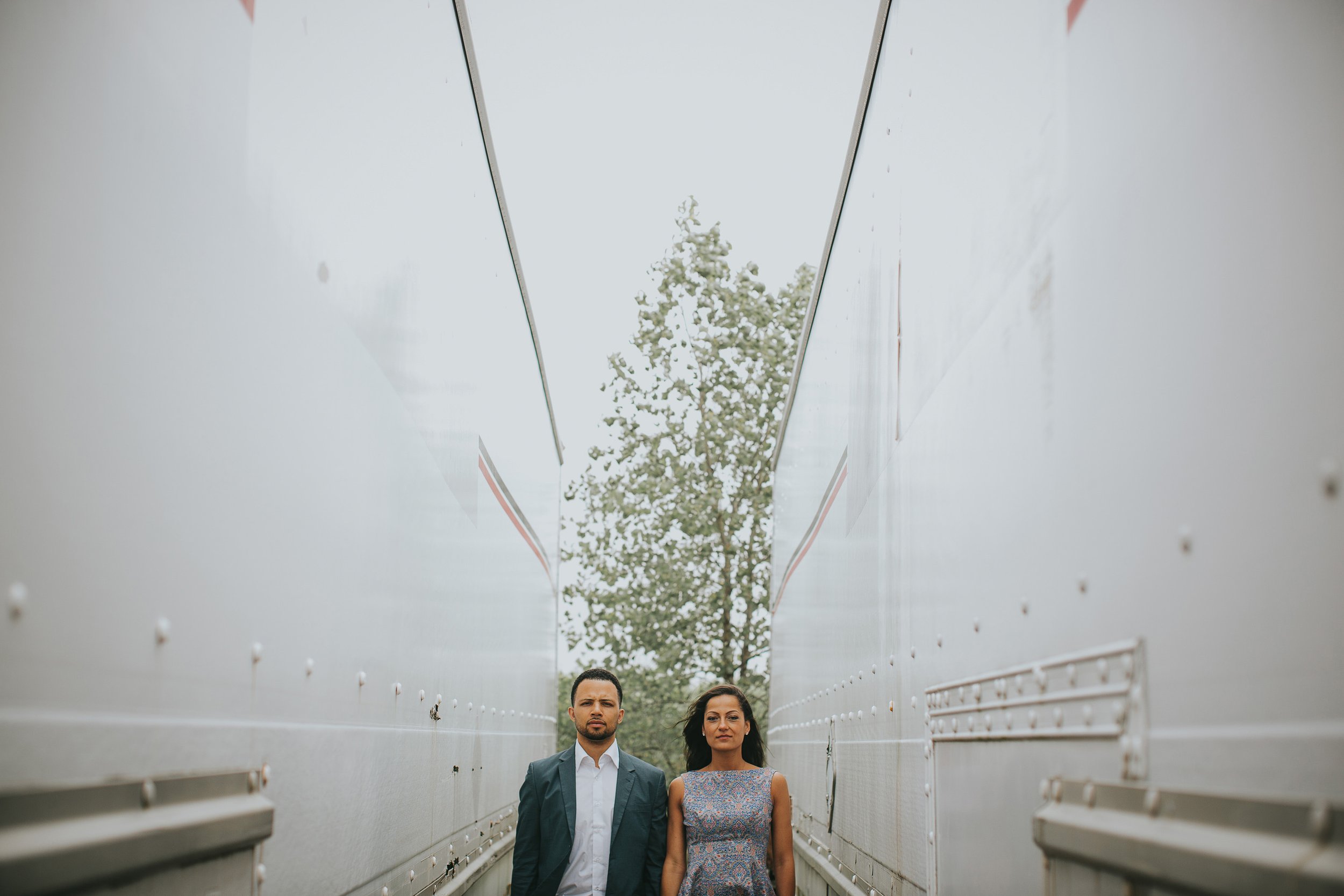 A couple stands together in an industrial setting, their serious expressions reflecting the strength and depth of their bond as they look forward to their future together.