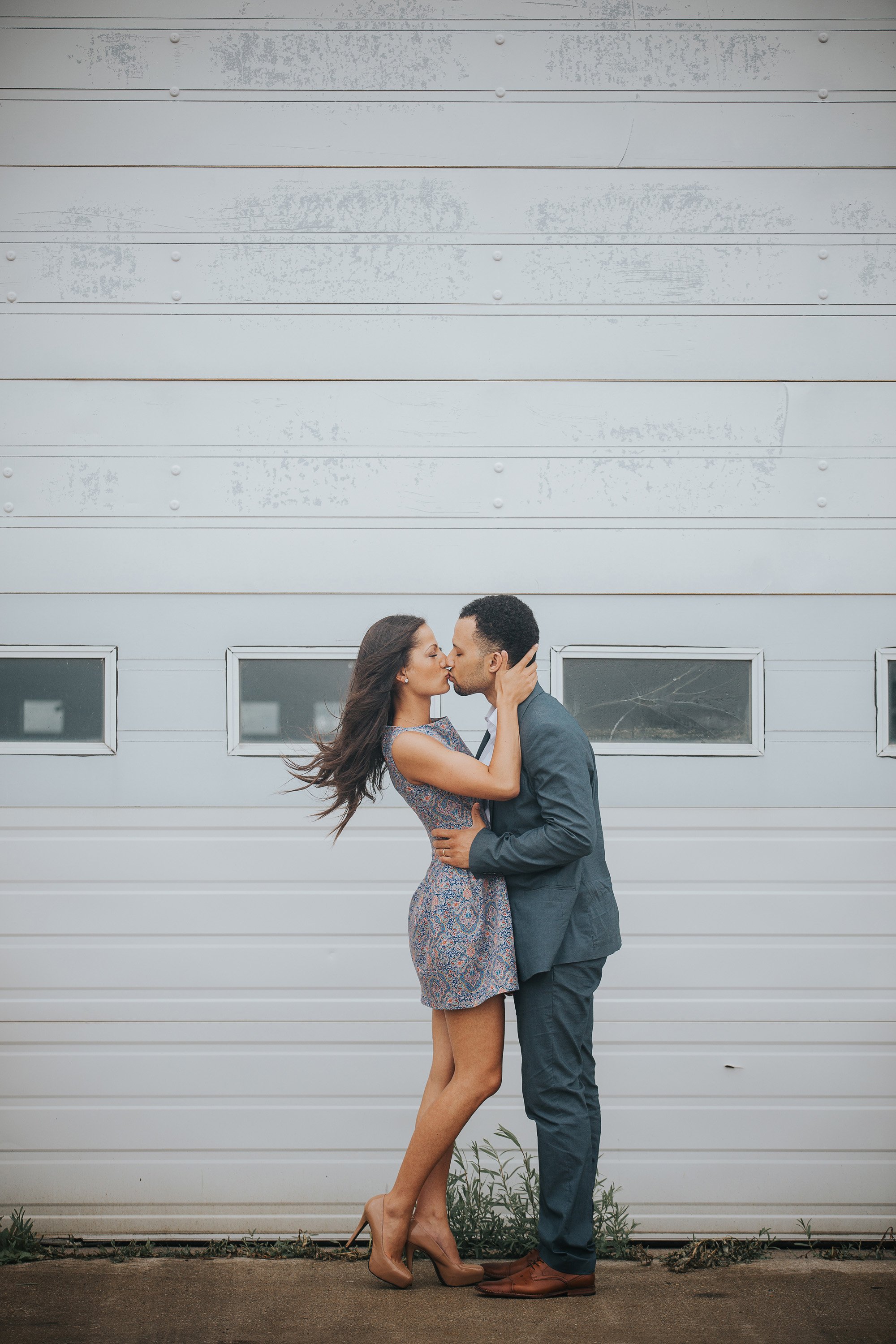 A couple shares a passionate kiss in an industrial setting, the close-up shot emphasizing their connection amidst the stark, textured background.
