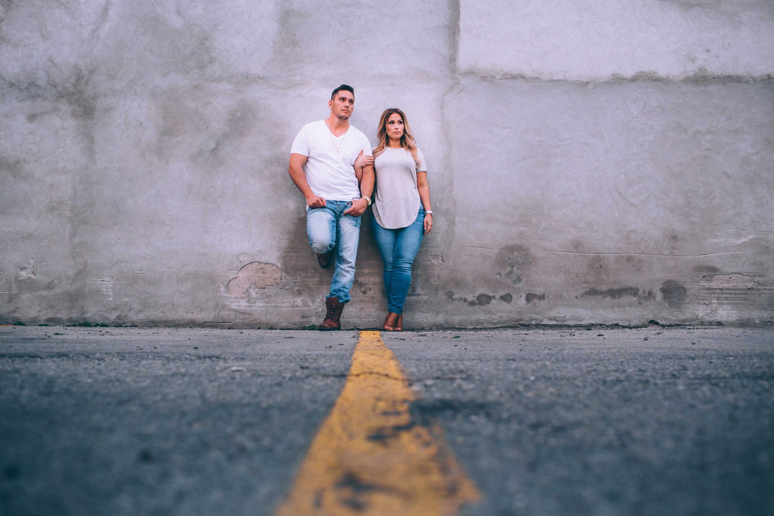 A casually dressed couple leans against an urban wall, posing together with a relaxed and confident demeanour.