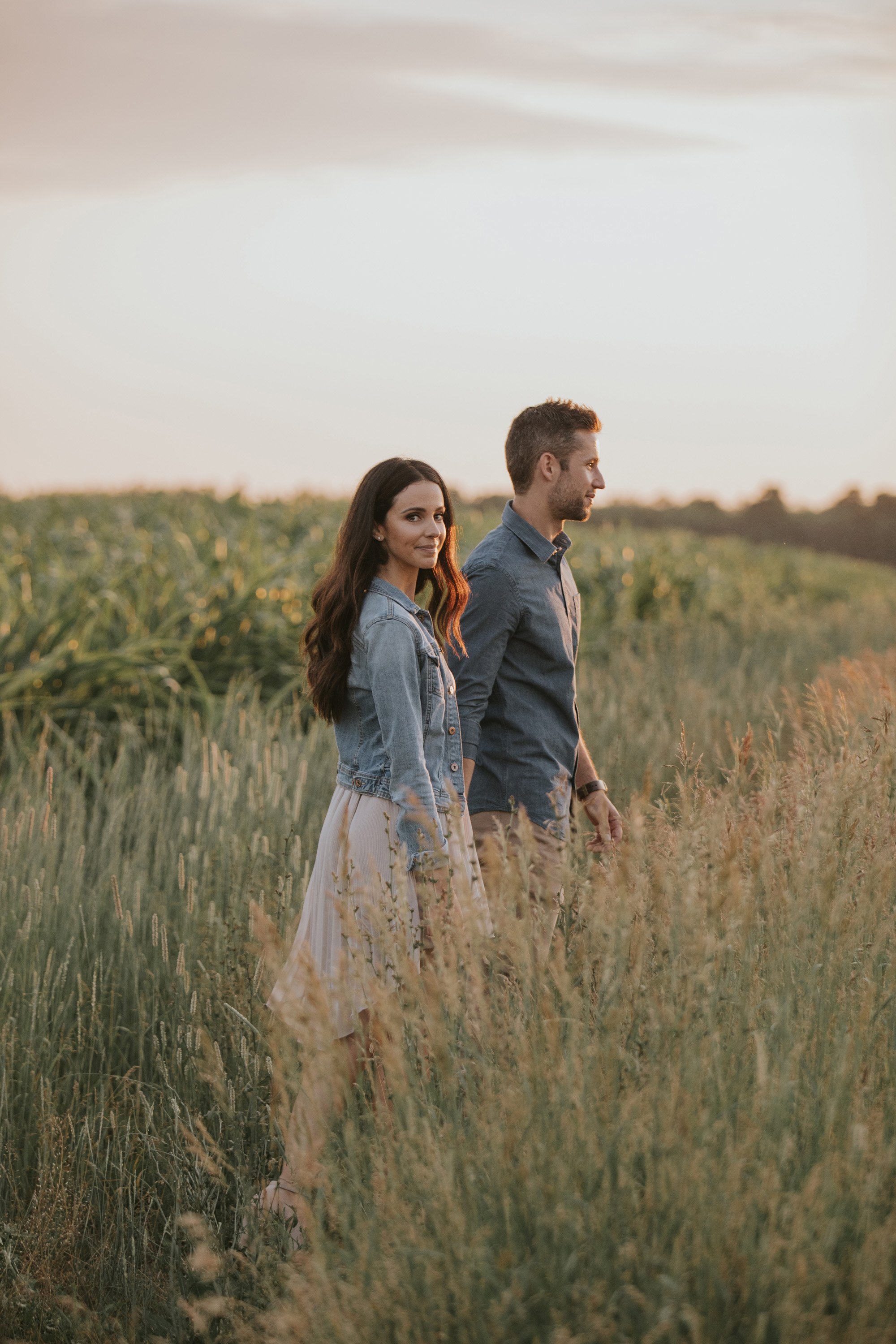 A couple walking through a sunlit field during sunset, both dressed casually with the woman wearing a light dress and denim jacket, and the man in a button-up shirt.
