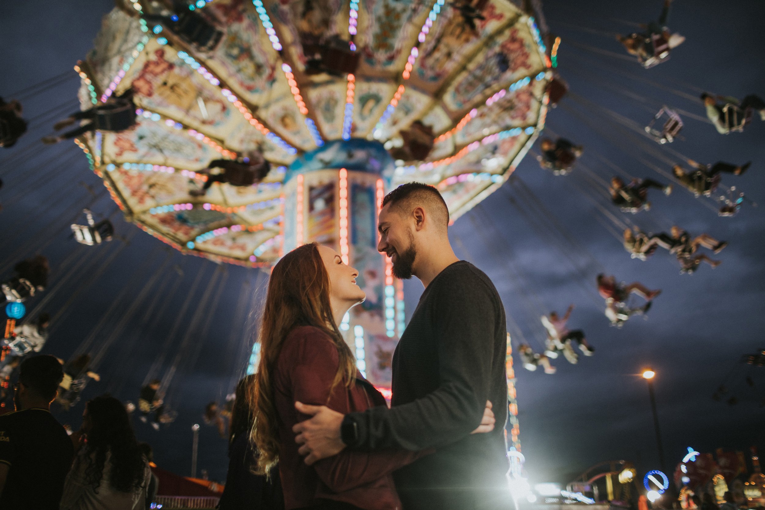 Couple standing close, smiling at each other under bright carnival lights, with a vibrant swing ride in the background at night.
