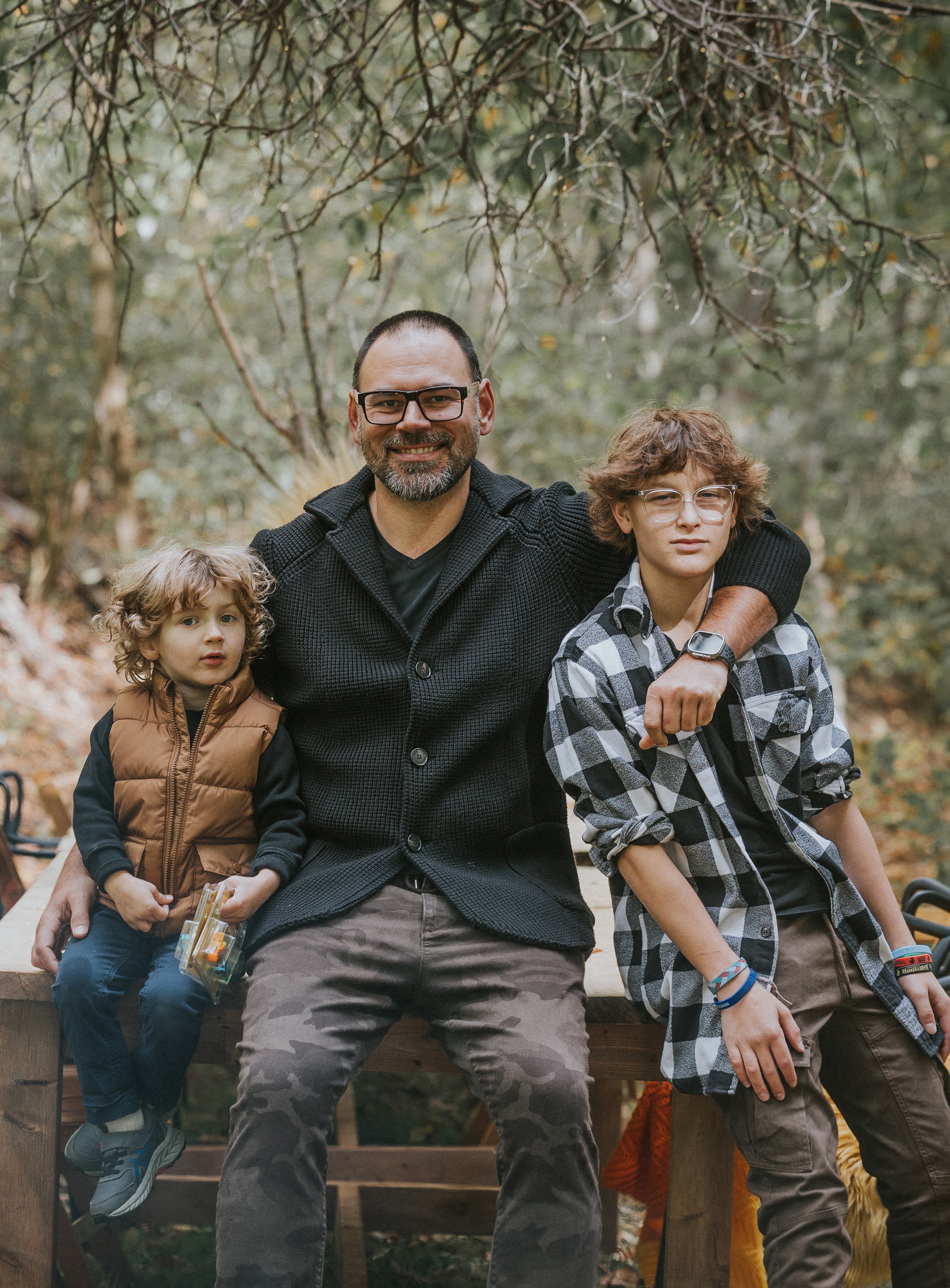 A father sits between his two sons on a wooden bench in an autumn setting, all smiling and casually dressed.