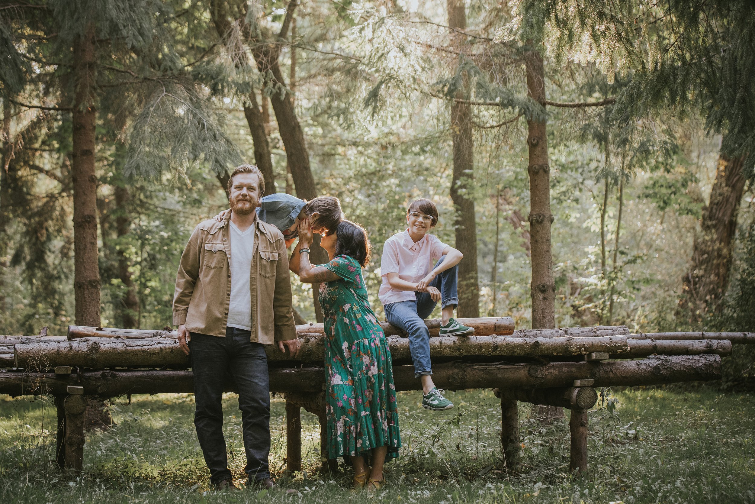 A family enjoying a playful and loving moment on a log bench in a sunlit forest, surrounded by tall trees.