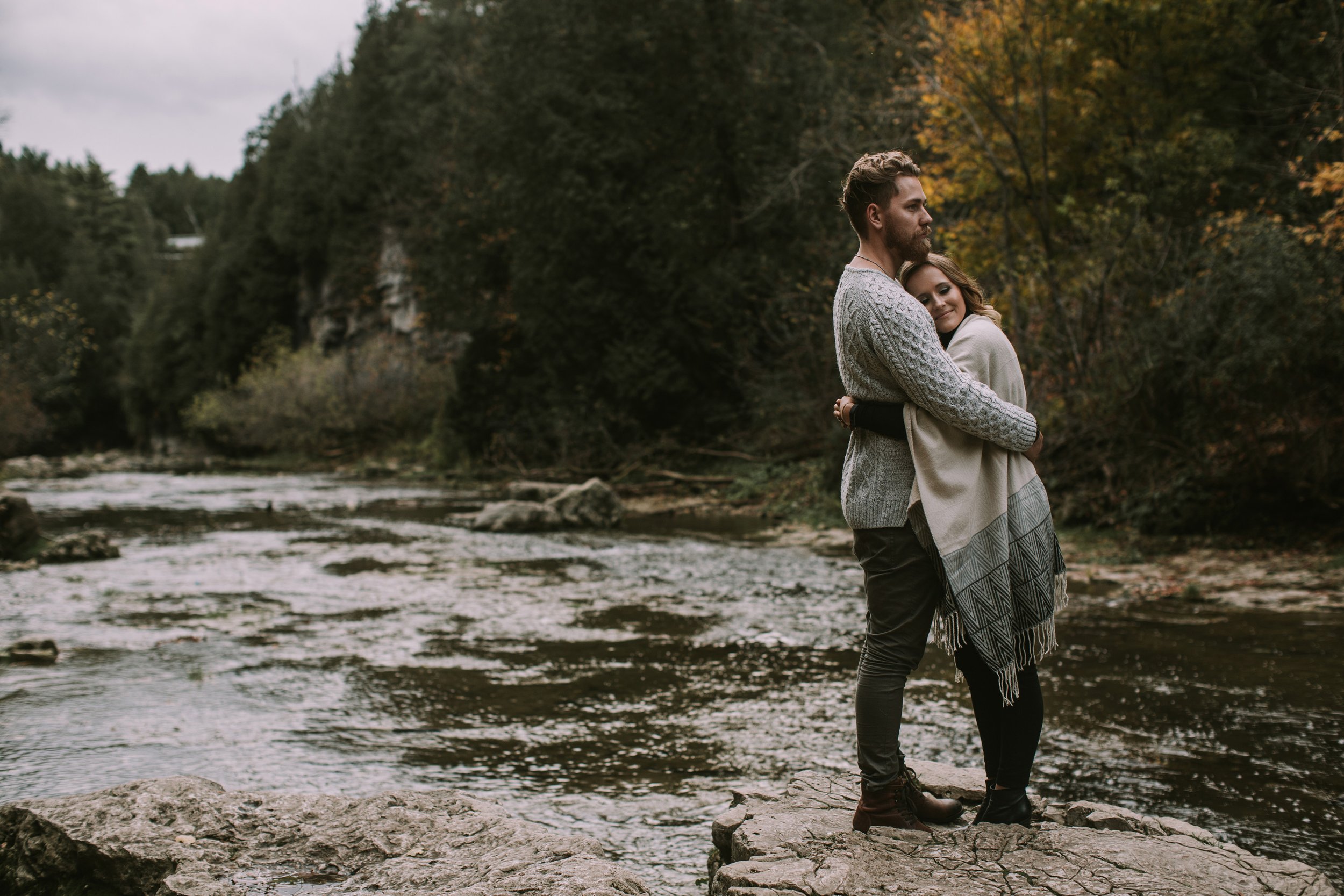 Couple embracing by a river in the fall, wrapped in cozy sweaters, capturing a moment of warmth and love.