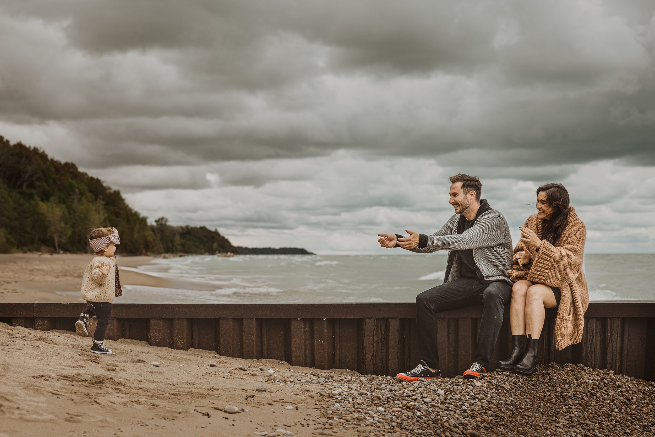 A joyful beach scene where a young daughter is running towards her parents, who are sitting on a wooden barrier, eagerly waiting with open arms. The cloudy sky and rolling waves in the background add a dramatic element to the playful family moment.