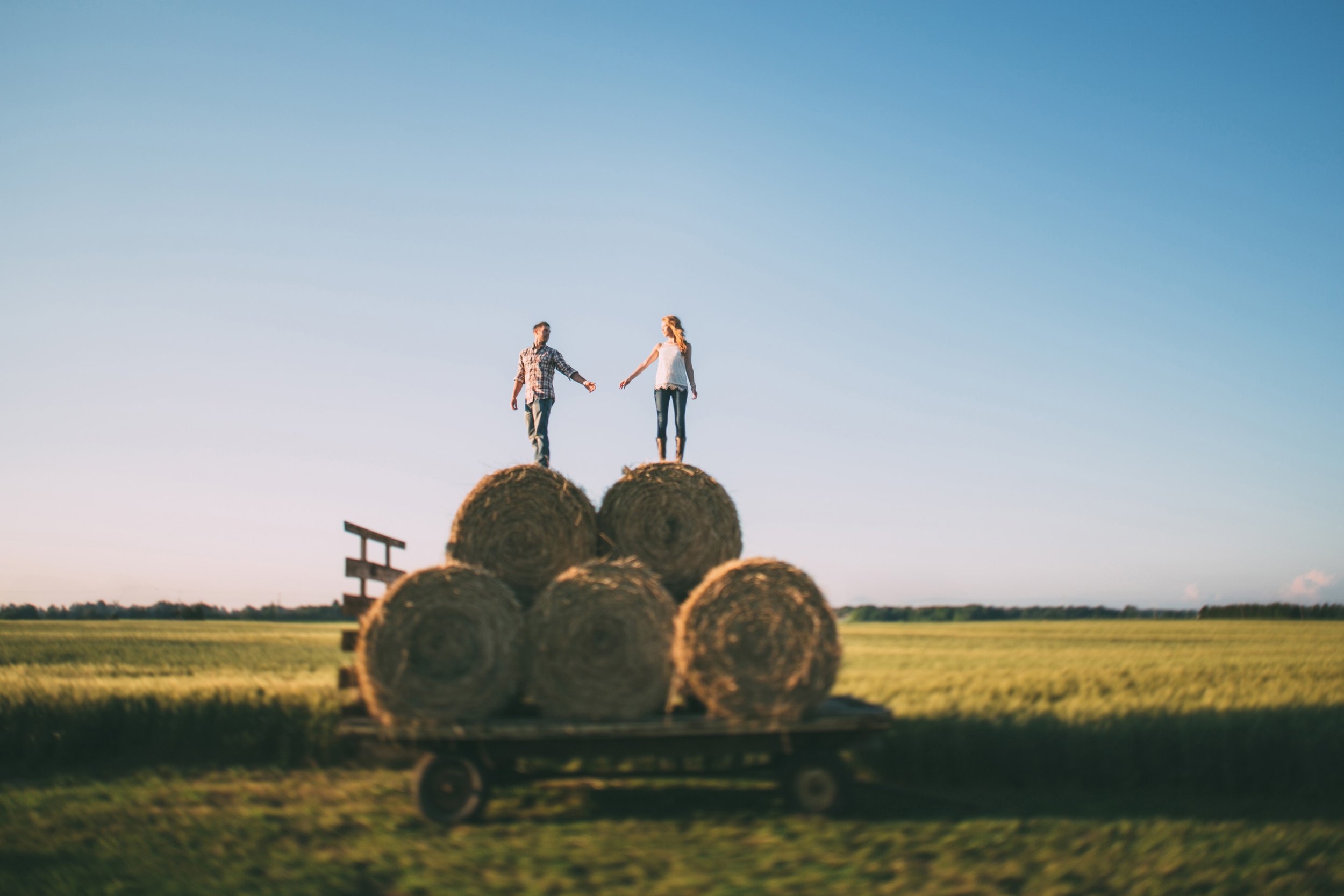 A playful engagement photo of a couple balancing on hay bales in a countryside field at sunset, capturing the joy and spontaneity of their relationship.