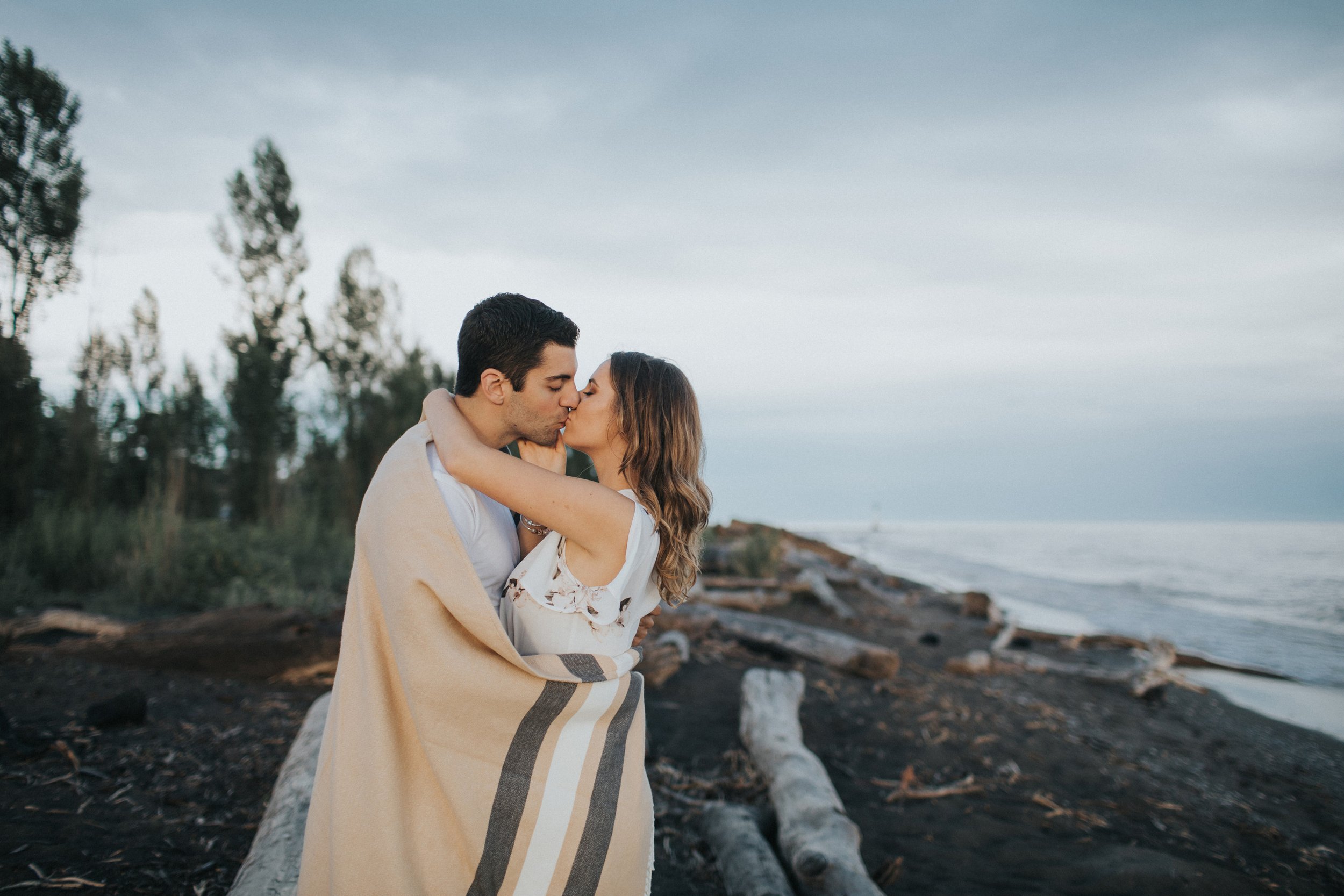 A couple shares a tender kiss wrapped in a blanket on a serene beach at sunset.