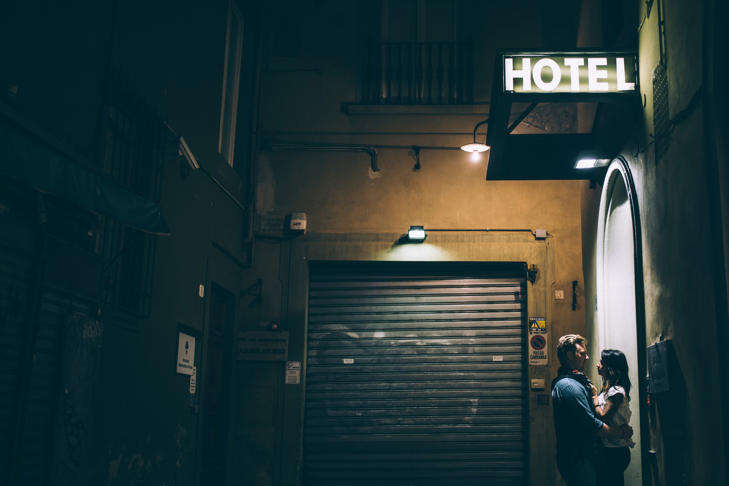 A couple embraces under the soft glow of a hotel sign, set against the backdrop of a dimly lit alley at night.
