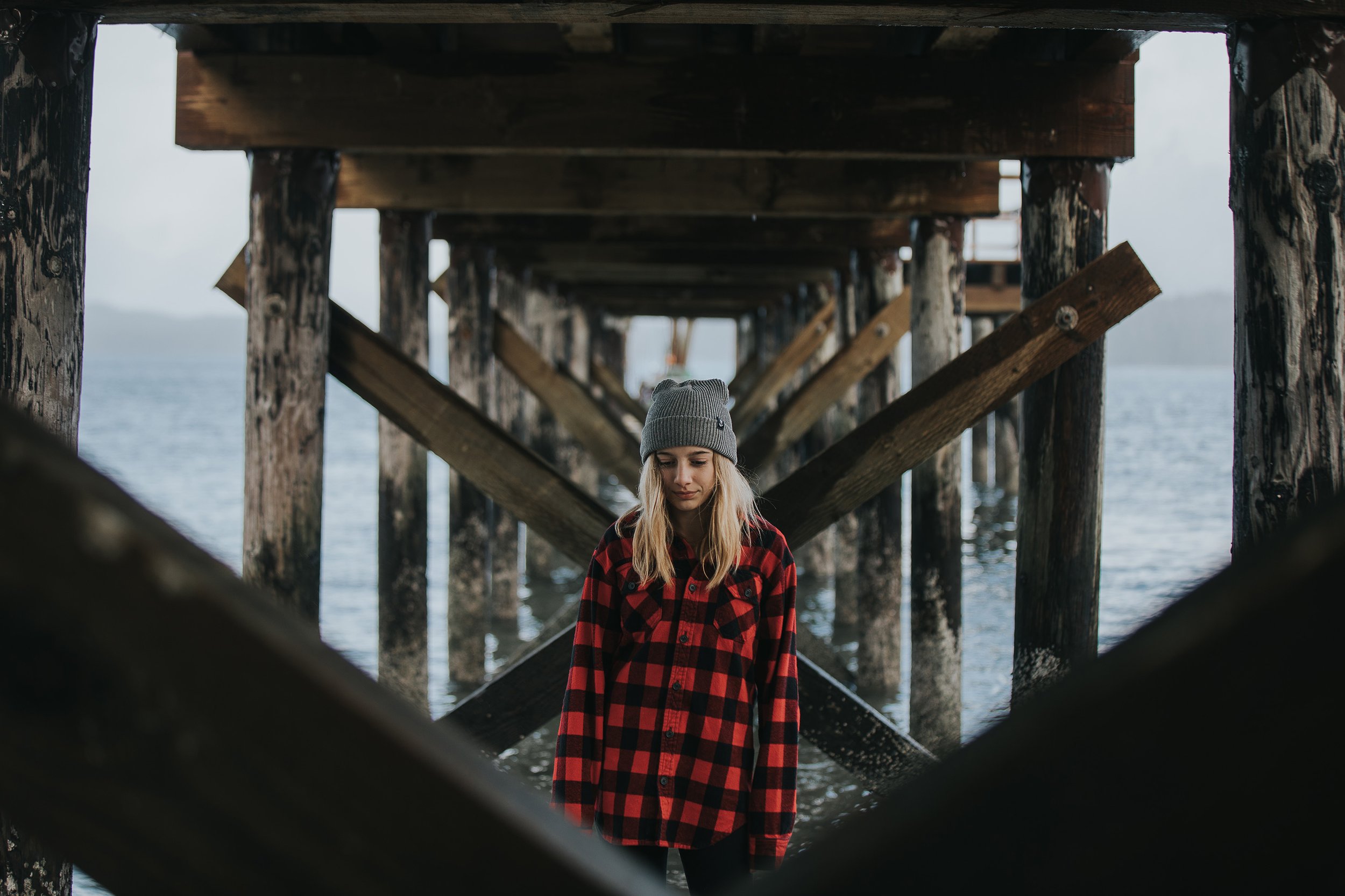 A girl in a red flannel shirt and beanie standing thoughtfully under a wooden pier, with the ocean behind her, capturing a moment of quiet adventure.