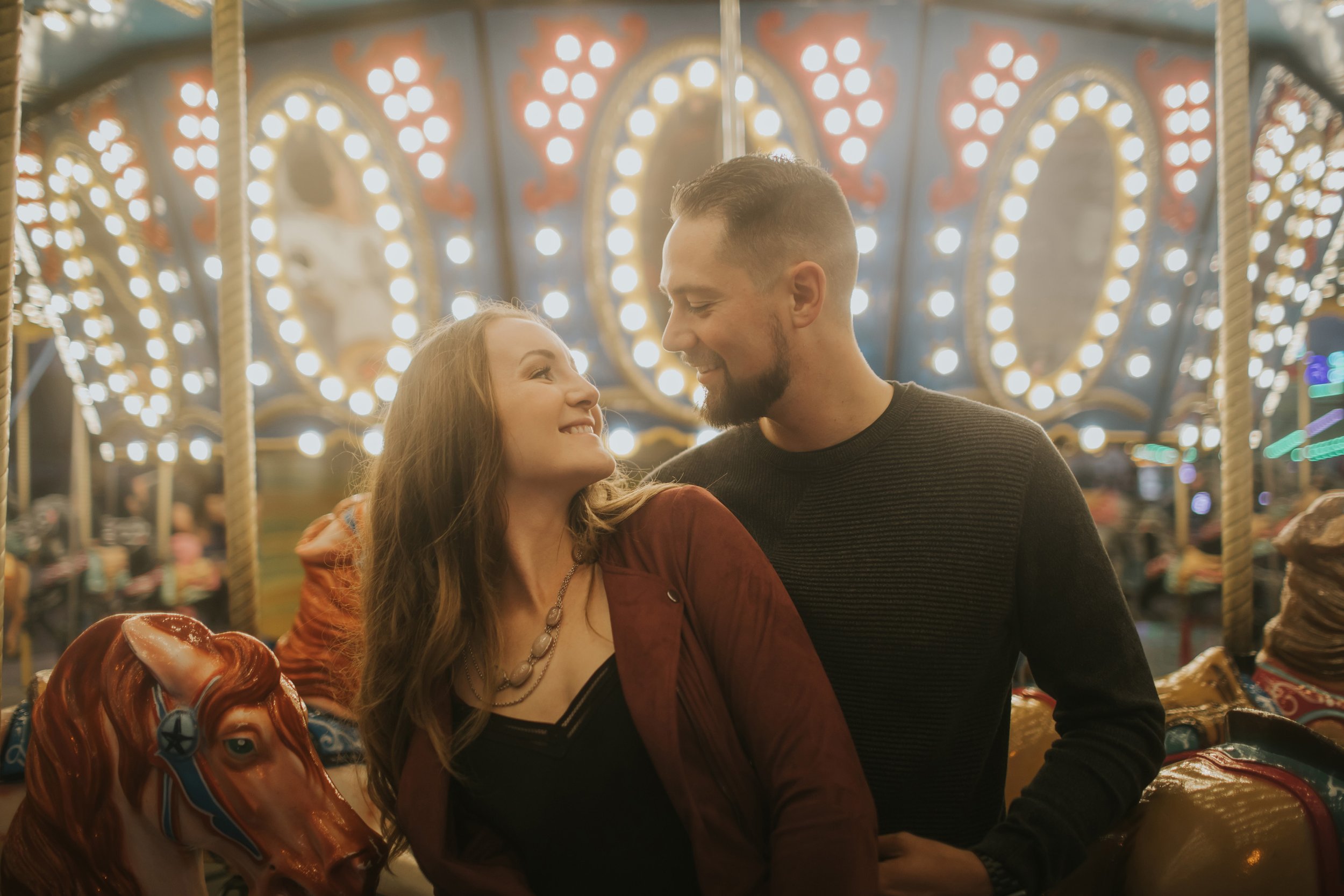 A couple enjoys a romantic moment together on a carousel ride, surrounded by vibrant lights and playful decor, capturing the joy of their engagement.