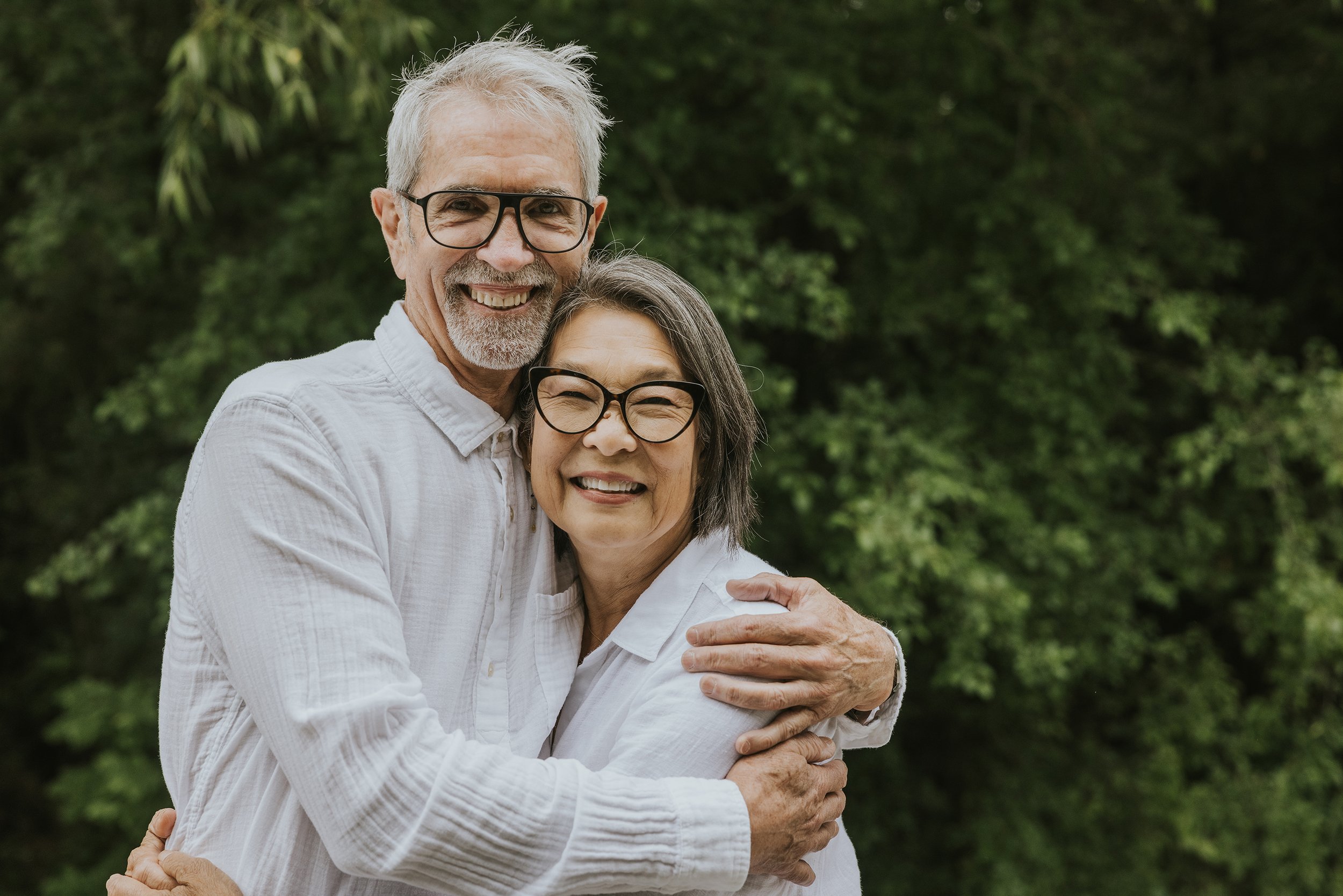 An older couple embracing each other with warm smiles in a lush, green park.