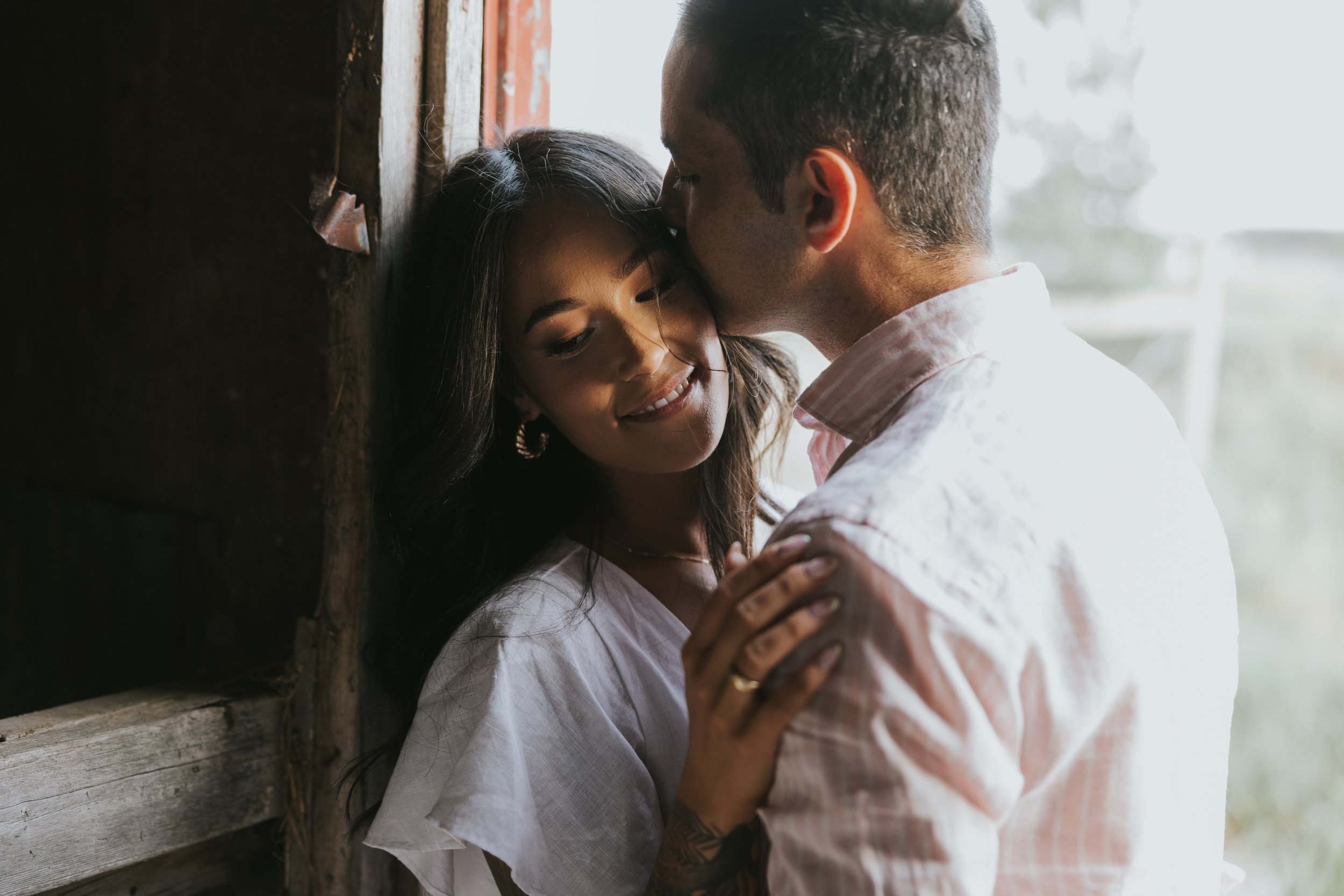 A couple shares an intimate kiss inside a rustic barn, with soft sunlight filtering through the wooden structure, capturing the warmth of their connection.