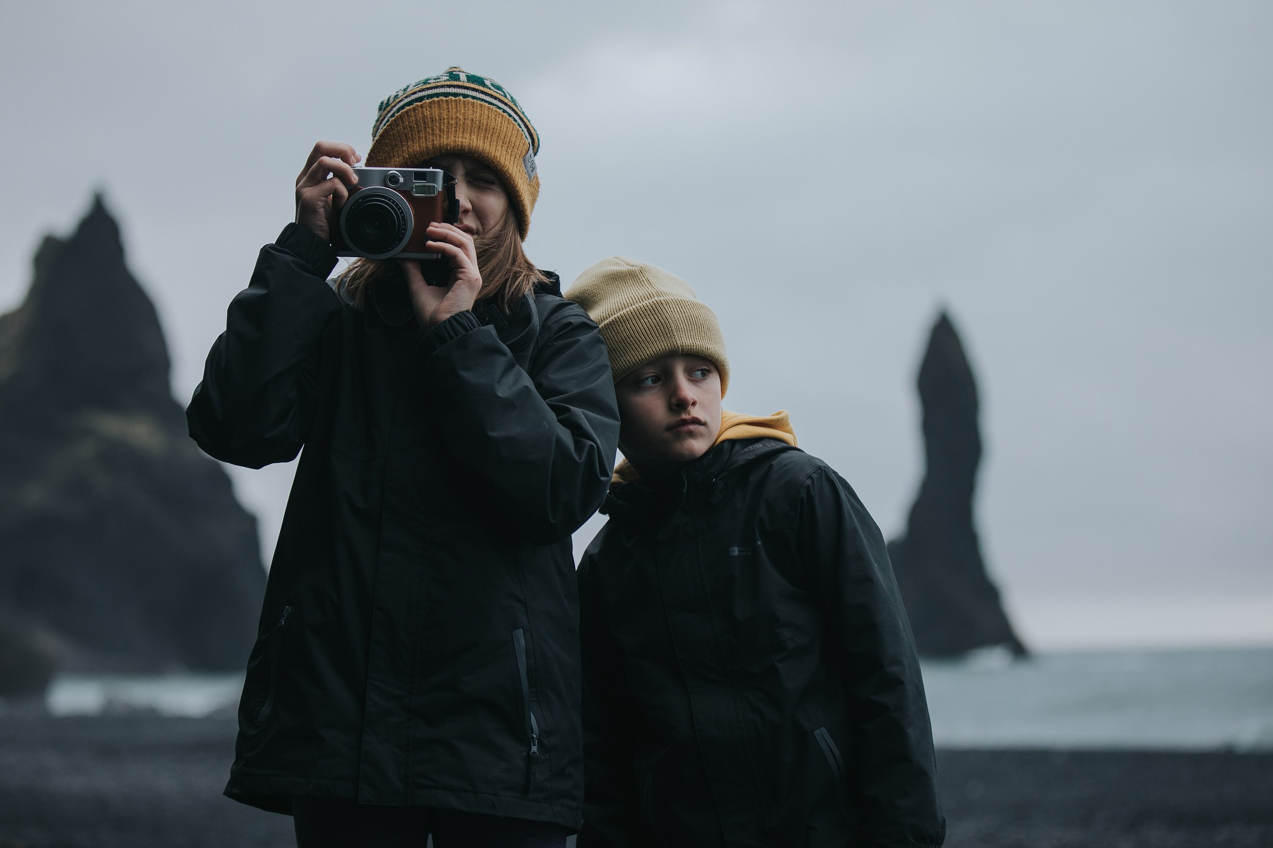 Two siblings dressed warmly, exploring a coastal area with rocky formations, one capturing a candid moment with a film camera.