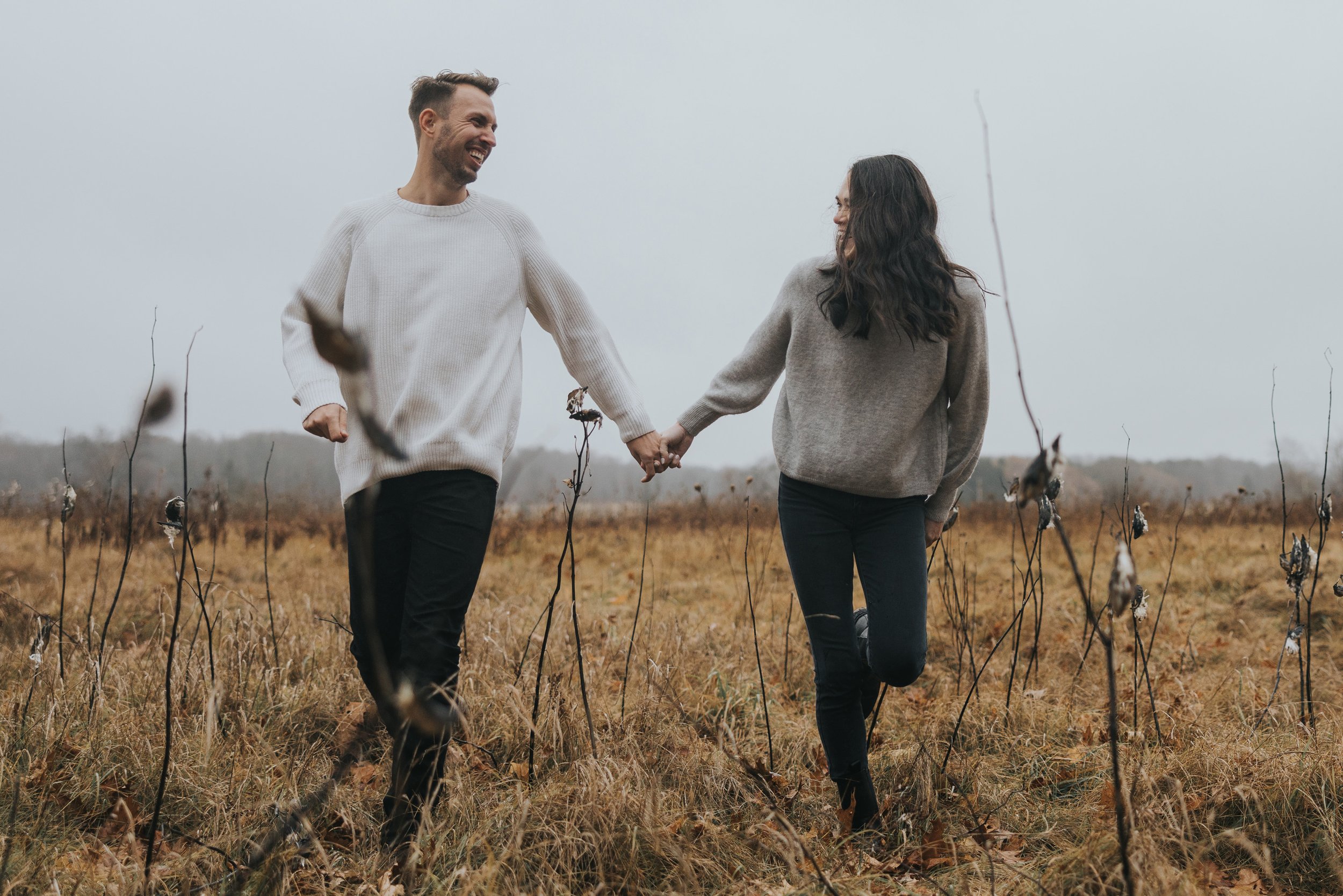 A couple runs hand in hand through an autumn field, laughing and enjoying their engagement session in London, Ontario.