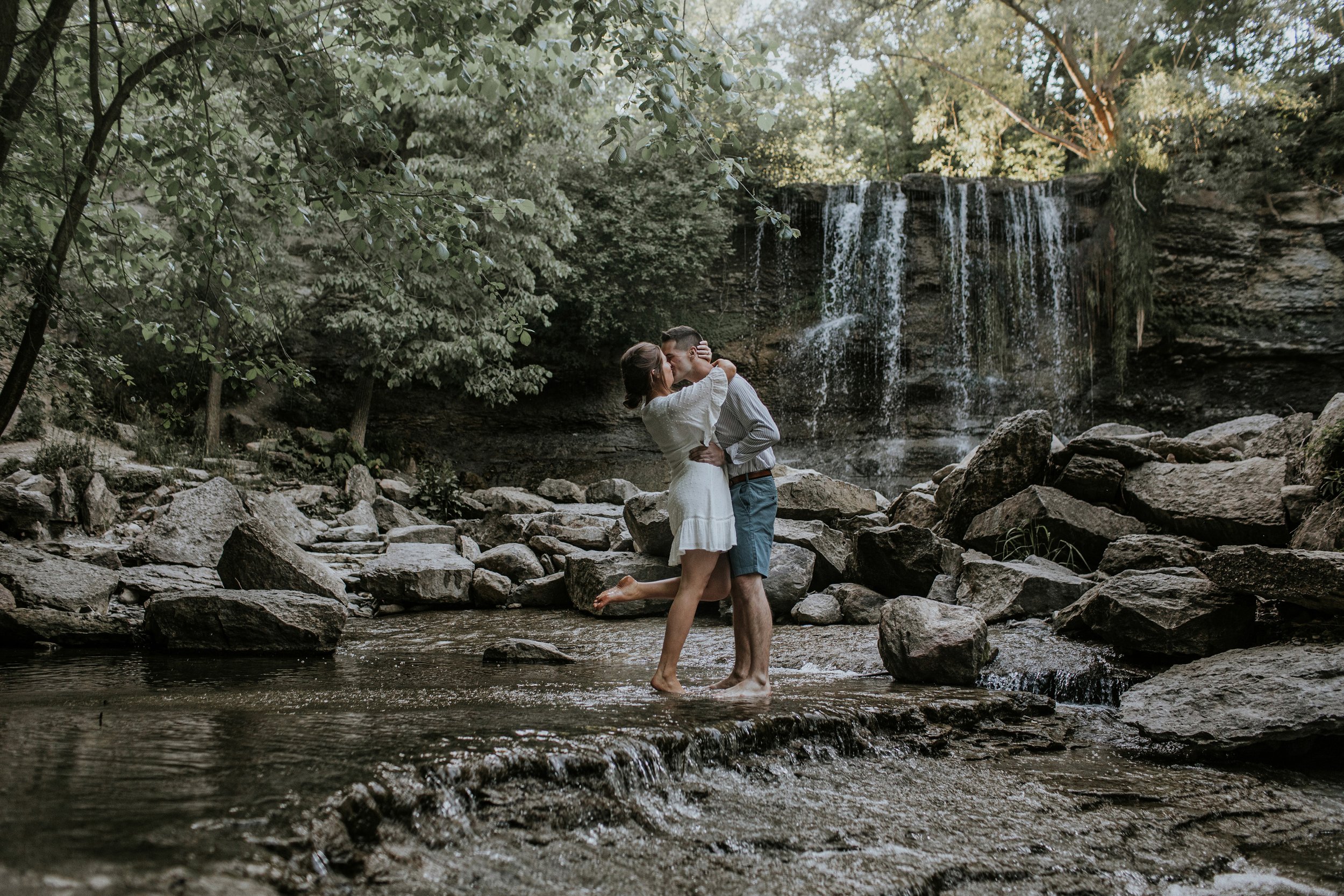 A couple embraces and shares a kiss while standing barefoot on rocks in front of a scenic waterfall, surrounded by lush greenery.