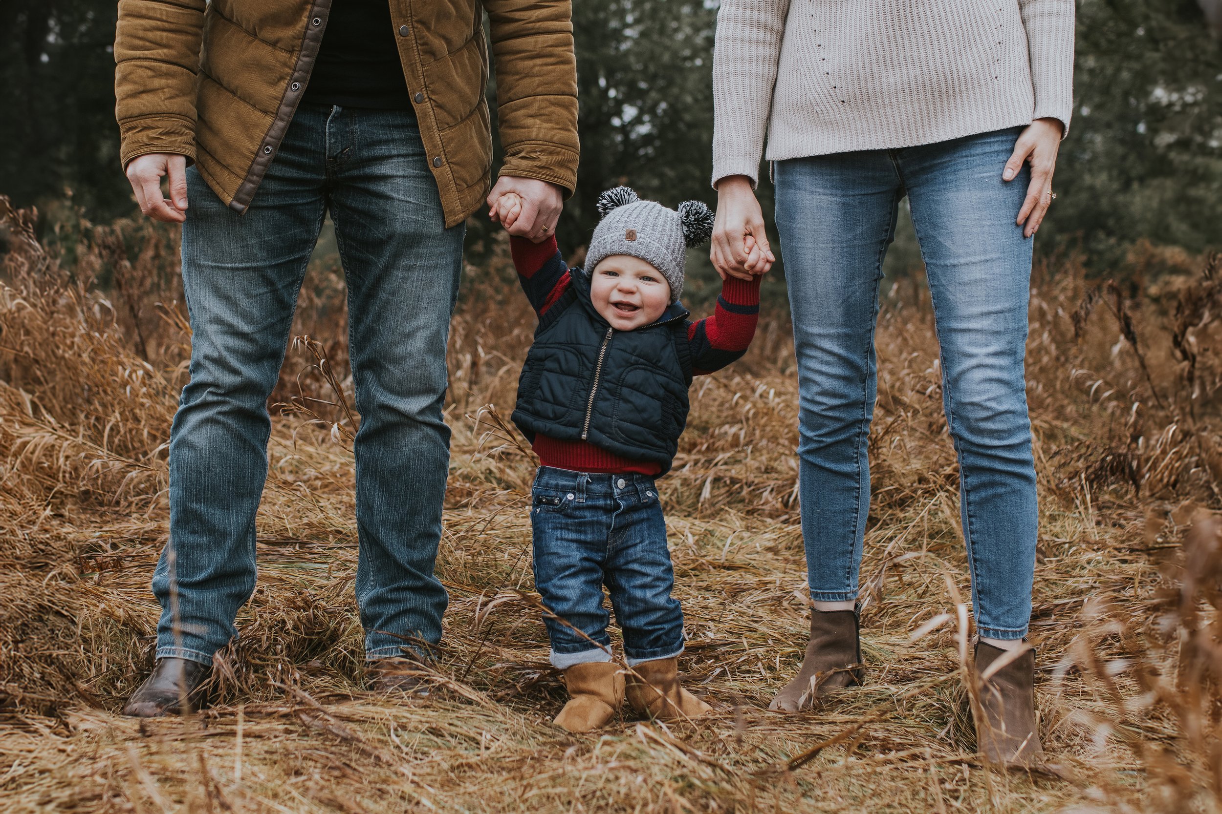 A baby holding hands with both parents while walking through an autumn field, dressed warmly and smiling brightly.