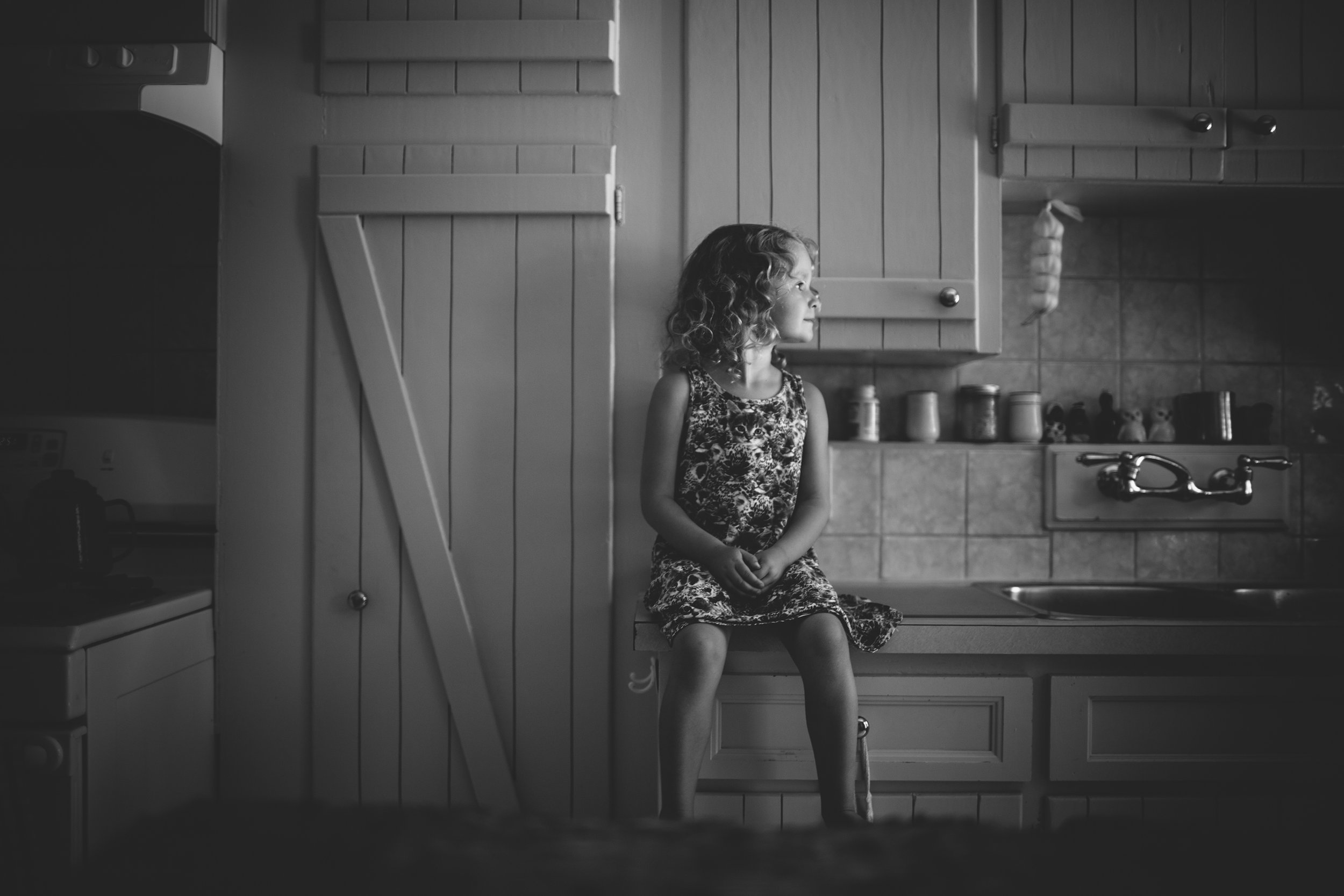 A young girl sits on a kitchen counter, bathed in natural light, in a kitchen with vintage decor, capturing a quiet, reflective moment.