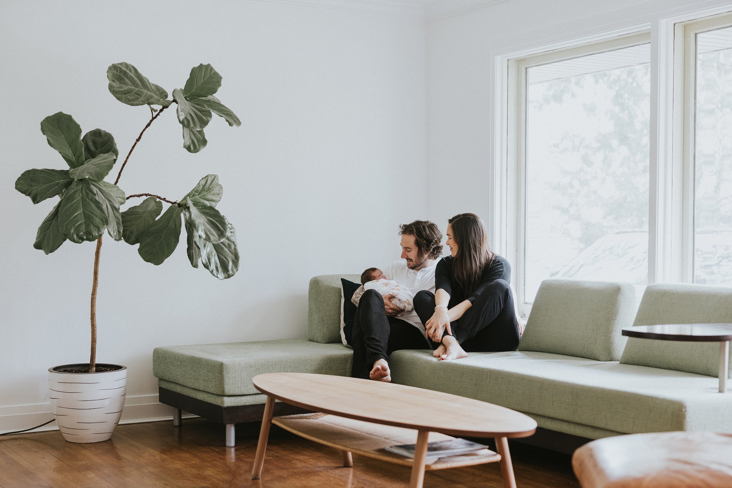 A family of three cuddling together on a modern green couch in a bright living room, sharing a peaceful moment with their newborn.