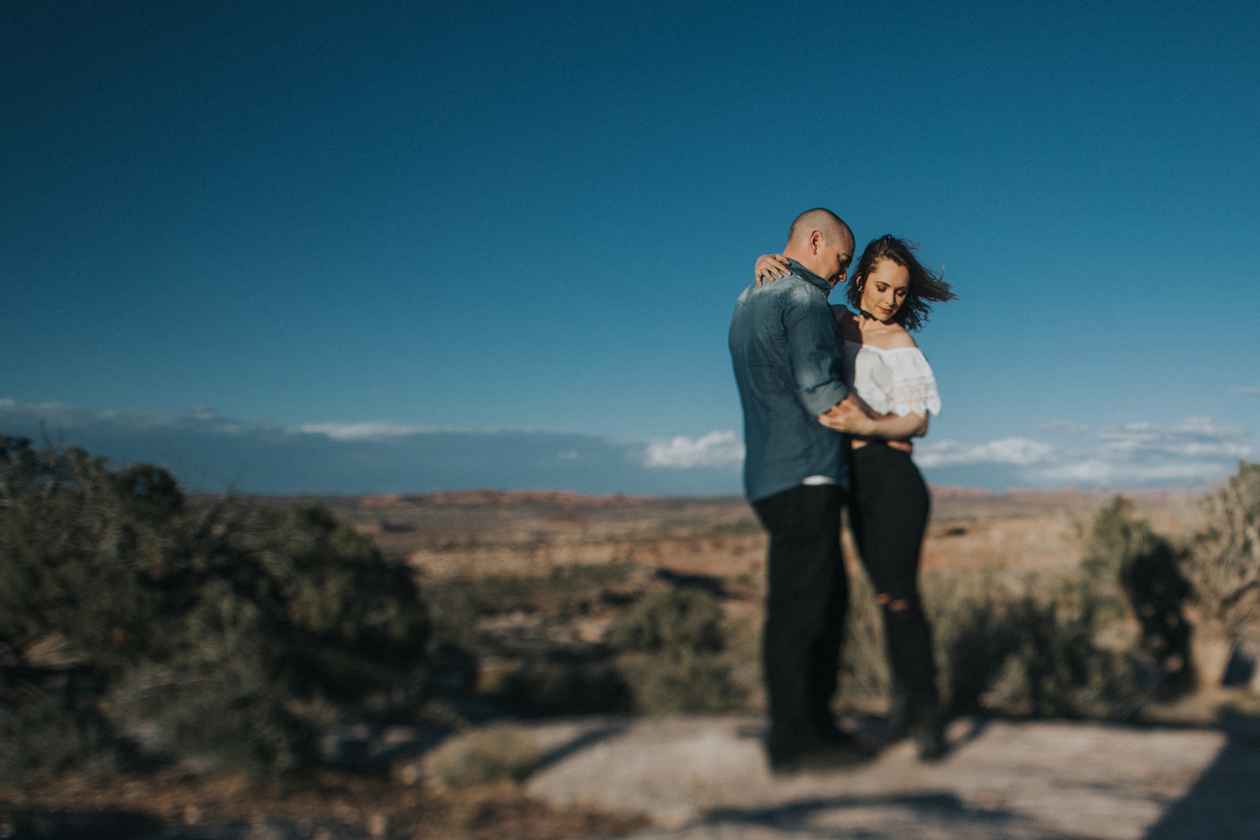 Couple standing close together in a desert landscape, surrounded by scenic views during their engagement photography session.