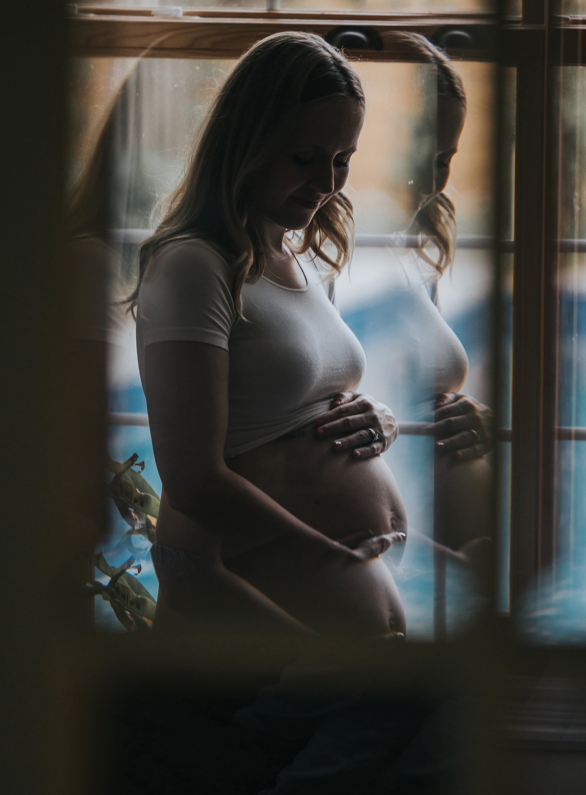 A tender maternity portrait of a woman gently holding her belly, with soft window lighting creating a serene reflection.