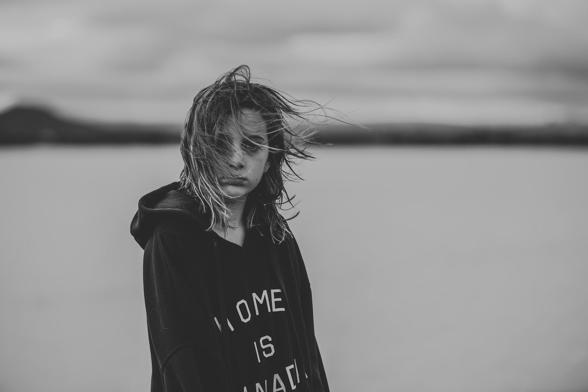 A black and white portrait of a child with windblown, wet hair standing near a serene body of water, embodying a sense of adventure.
