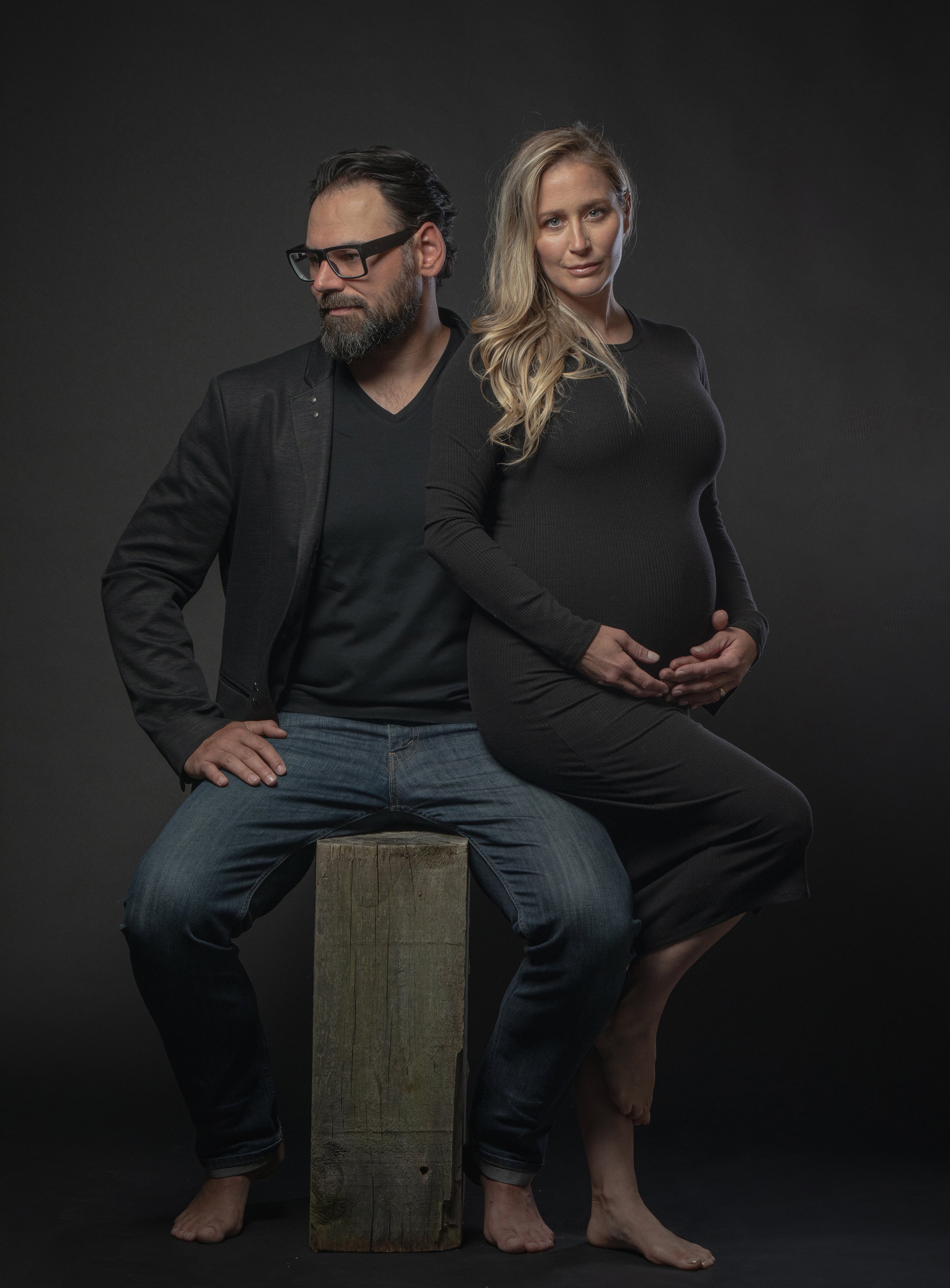 A dramatic studio portrait of an expecting couple, with the man sitting on a wooden stool and the woman standing behind, cradling her baby bump.