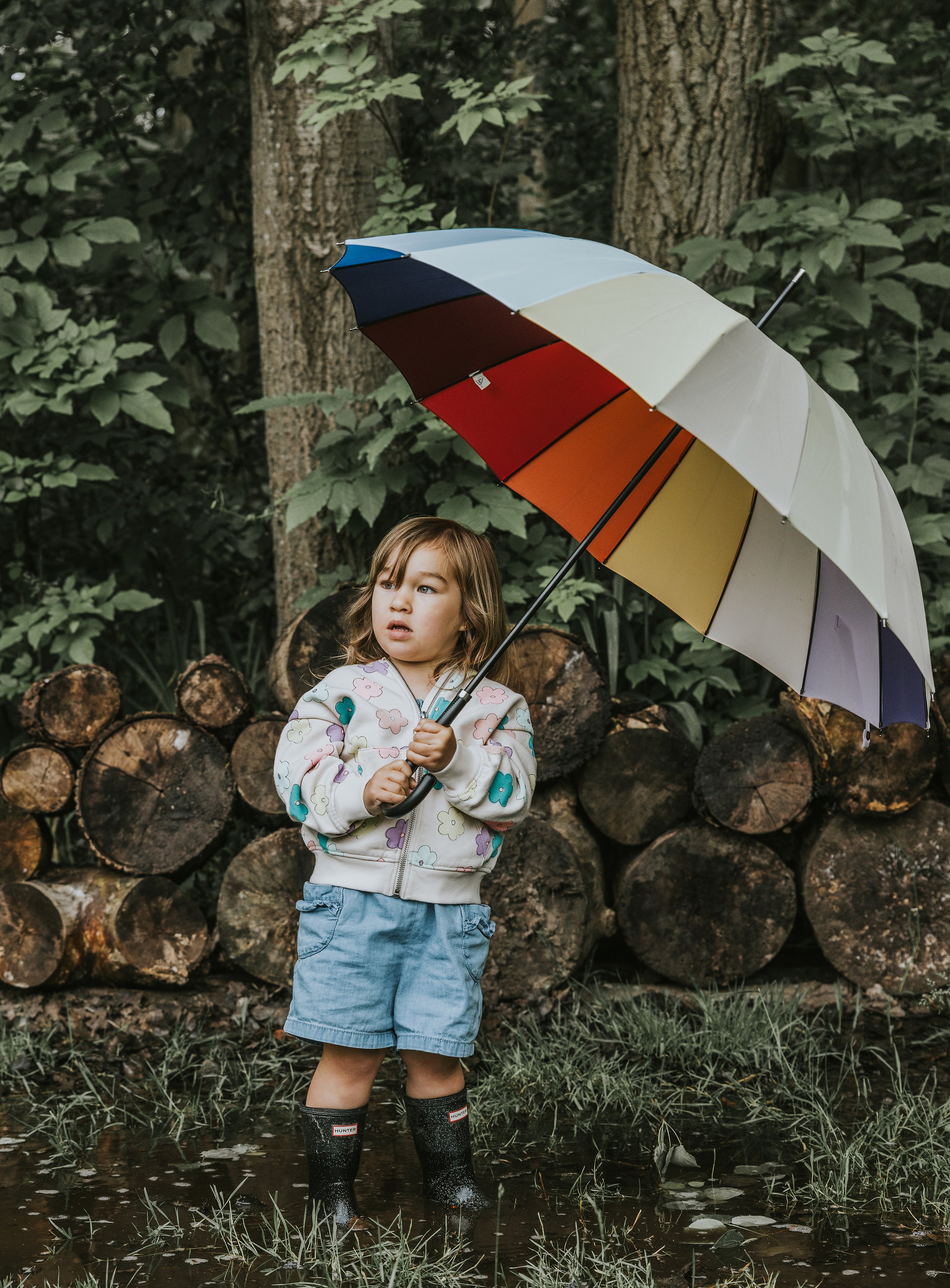 Adorable young girl standing outdoors, holding a colourful umbrella, with a backdrop of stacked logs and greenery.