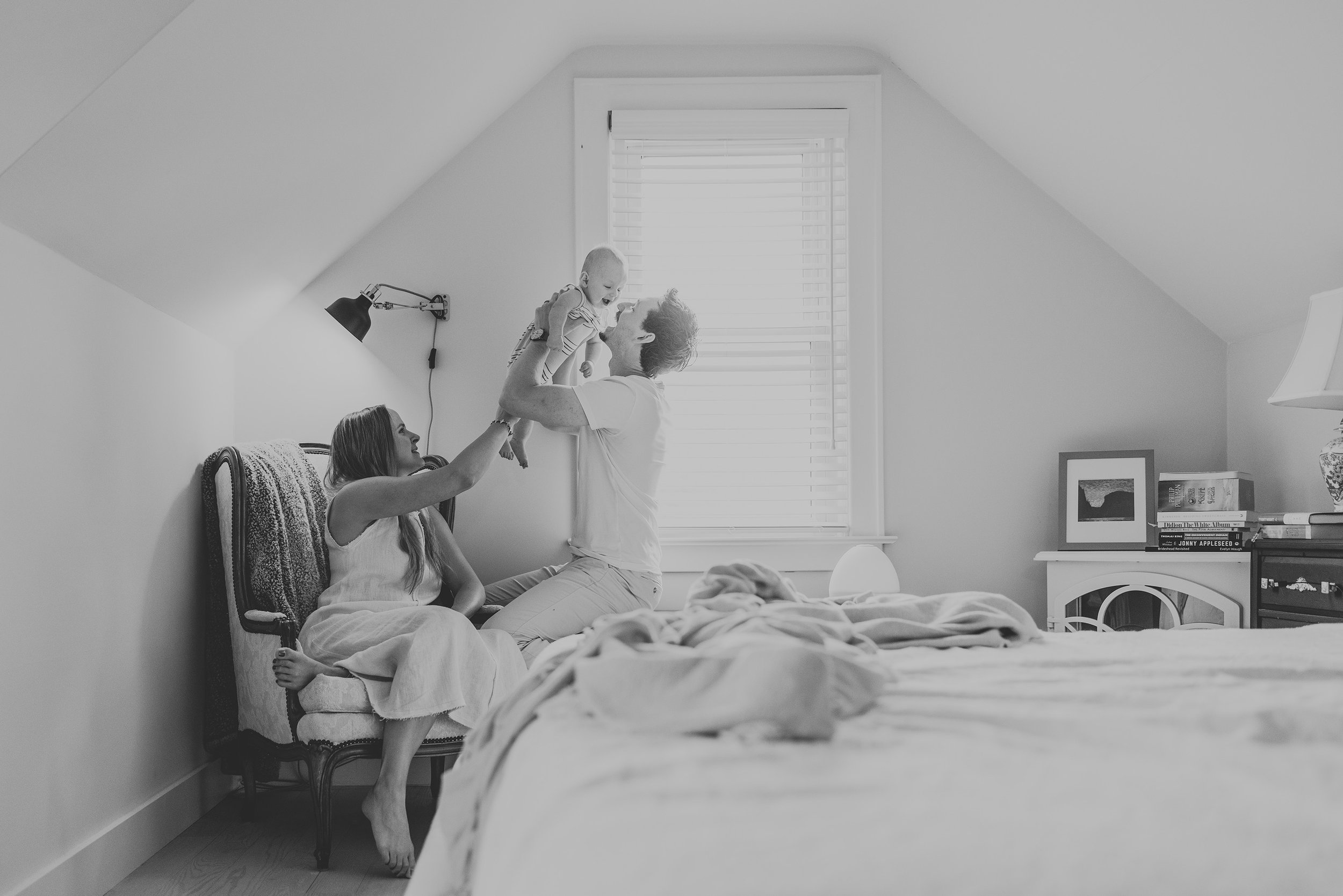 A candid family moment in a bedroom, with parents playing with their baby in a cozy, light-filled space.