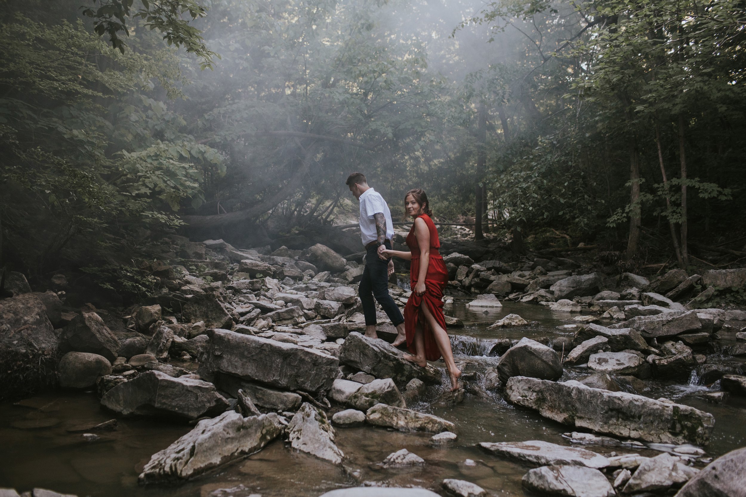 A couple walking hand in hand across a rocky riverbed, with the woman in a red dress leading the way, creating a romantic and adventurous atmosphere in a natural setting.