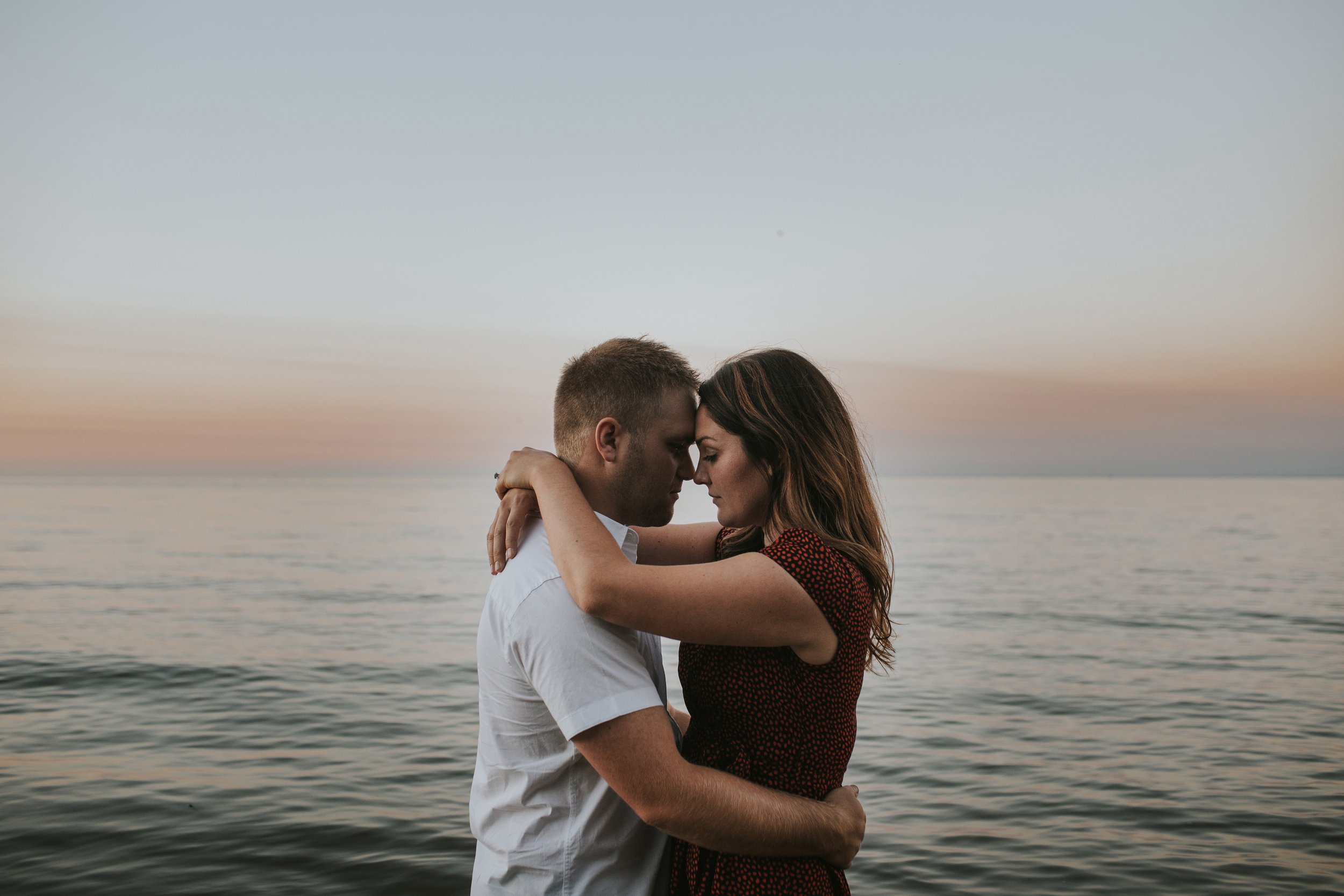 A couple embraces by the lake at sunset, the calm waters and warm colors of the sky creating a serene and intimate moment.