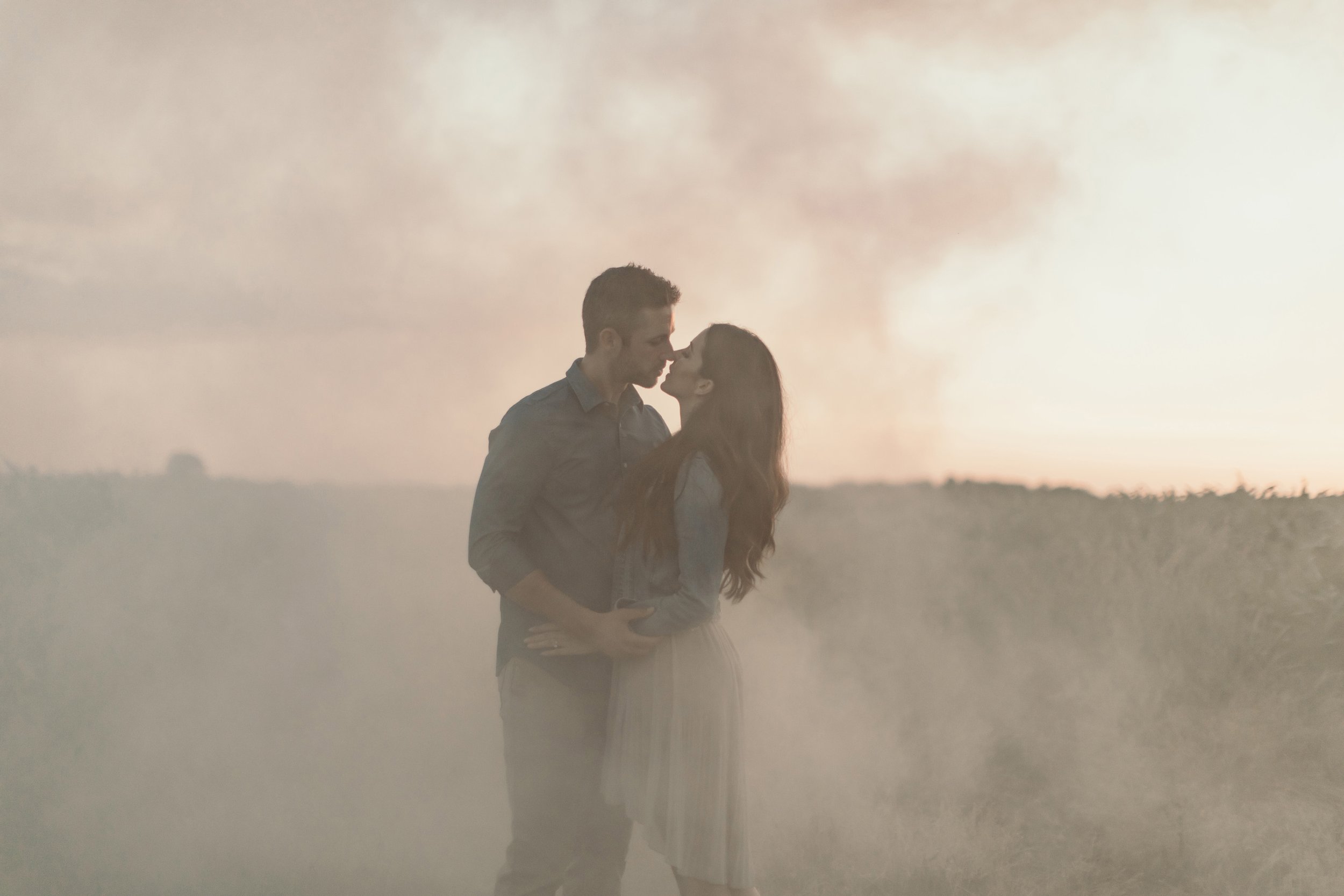 Couple sharing a kiss in a misty field at sunset, with a soft glow of light filtering through the haze.