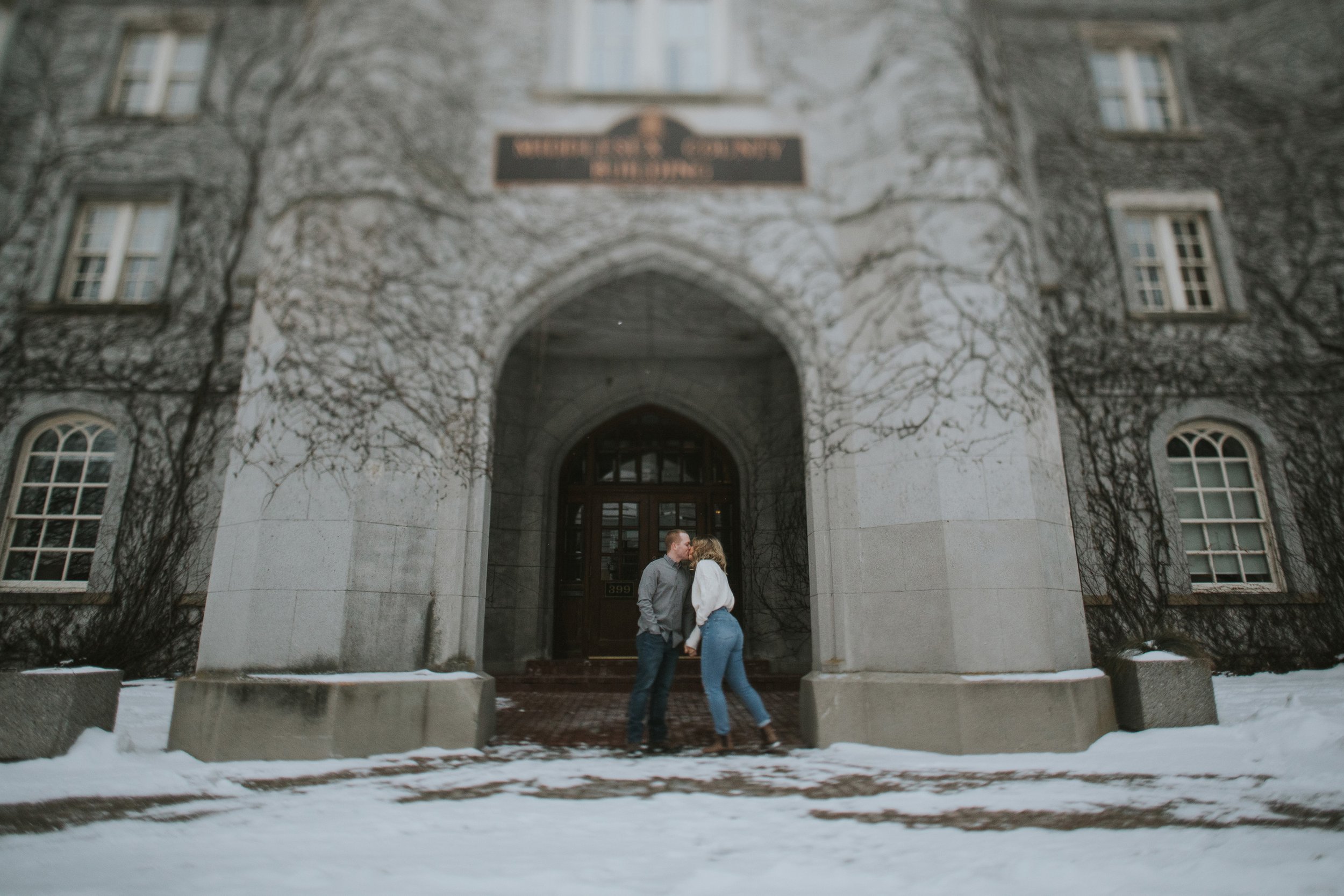 A couple shares a kiss in front of a historic building during winter, with snow on the ground adding to the cozy and nostalgic atmosphere of their engagement.