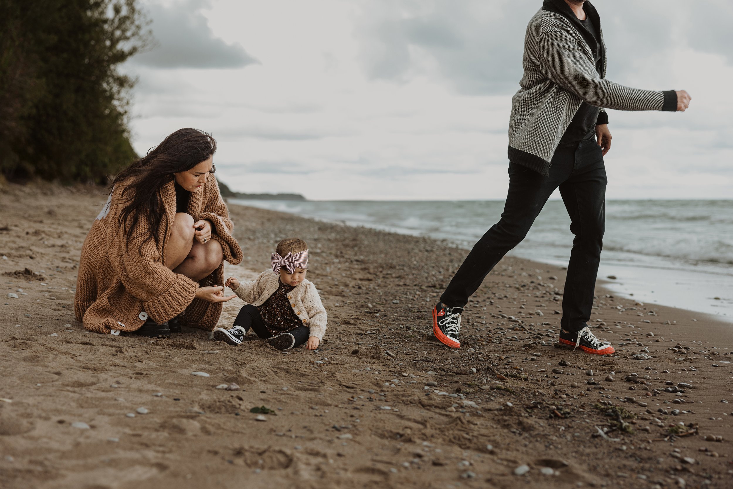 A family moment at the beach where a mother is crouched down, holding her young daughter's hand as they explore the sand together. The father is seen walking away in the background, adding a dynamic element to the peaceful, overcast beach setting.