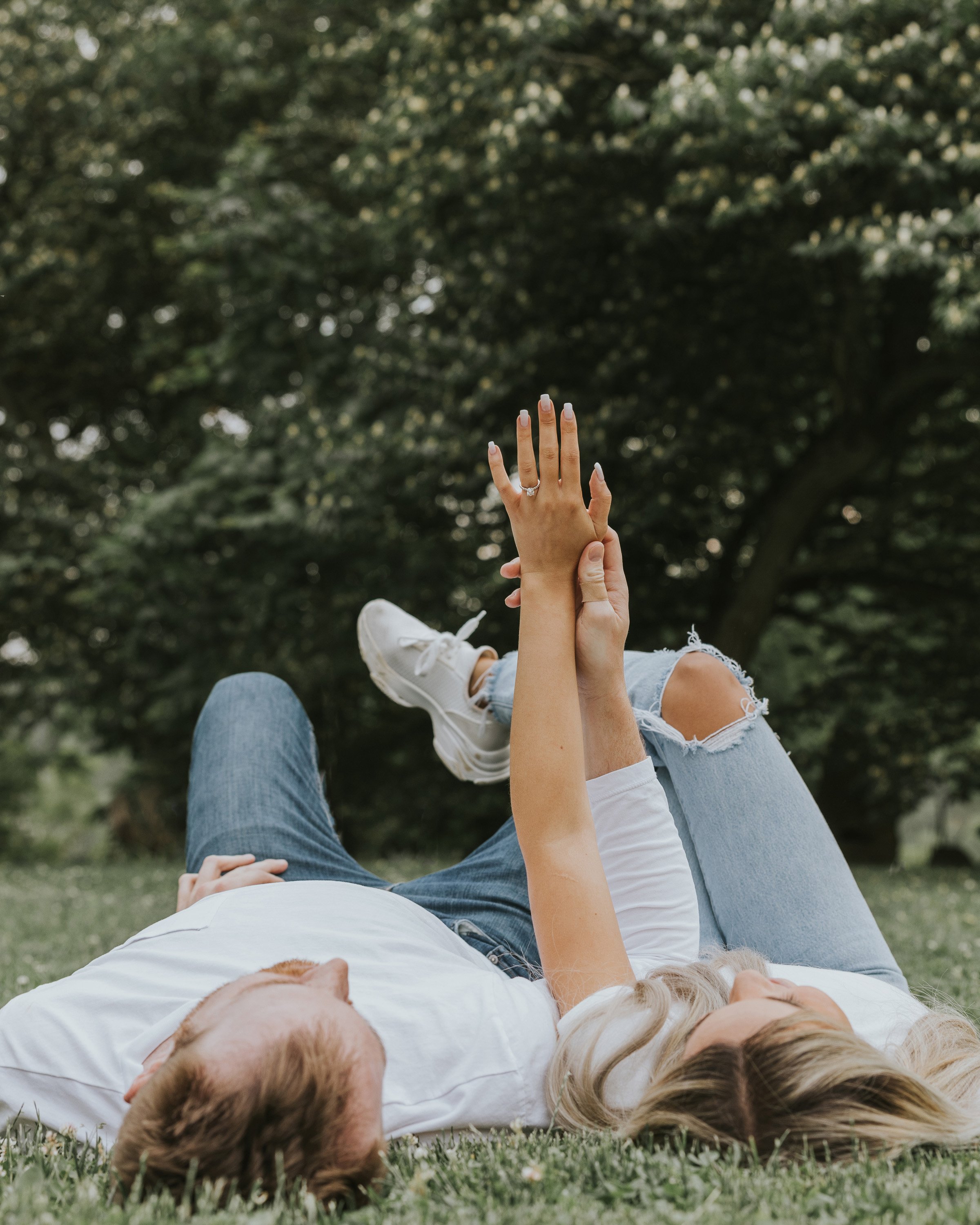 A couple lying on the grass, holding hands up towards the sky, enjoying a peaceful moment surrounded by nature.