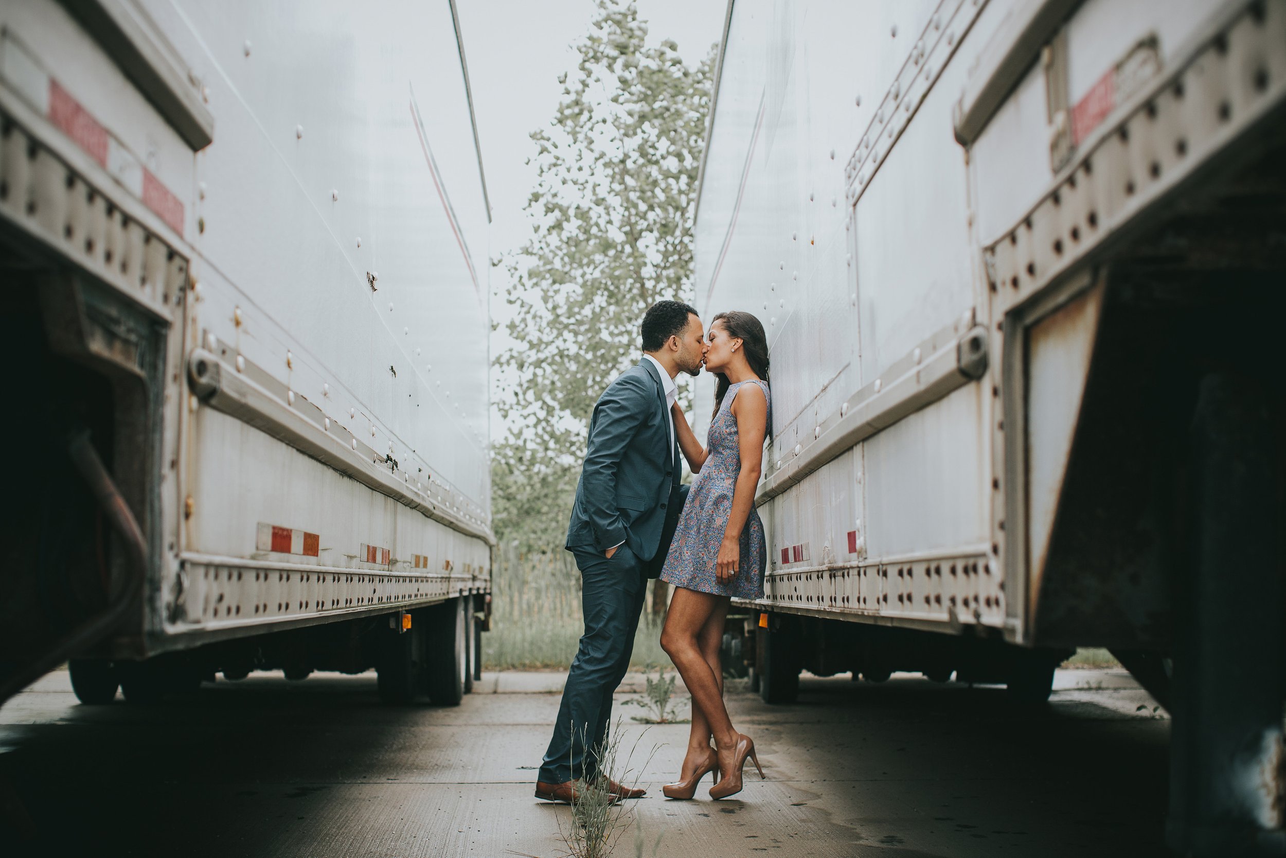 A couple shares a kiss between two large trucks in an urban setting, creating a unique romantic atmosphere.