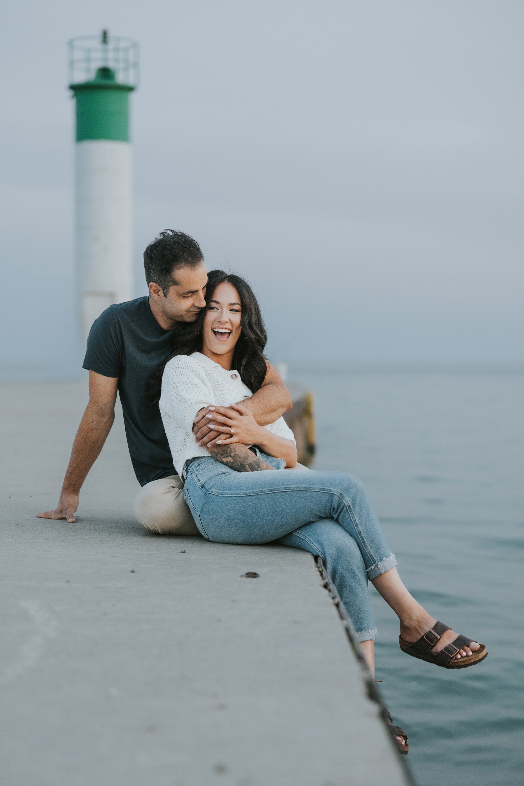 A couple sits on a pier near a lighthouse, sharing a loving connection with serene water in the background.
