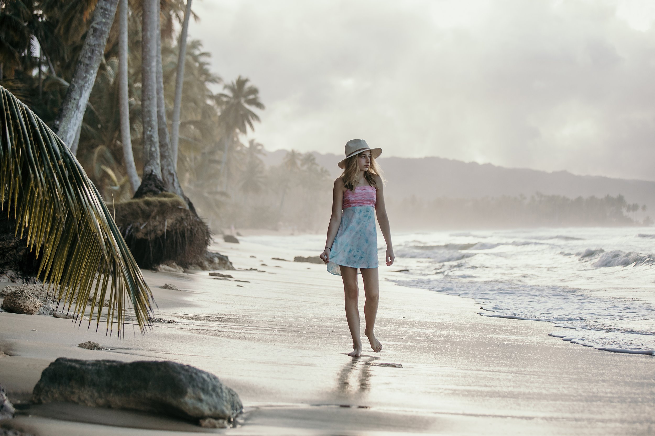 A young girl walking barefoot along a tropical beach at sunset, with palm trees and waves in the background.