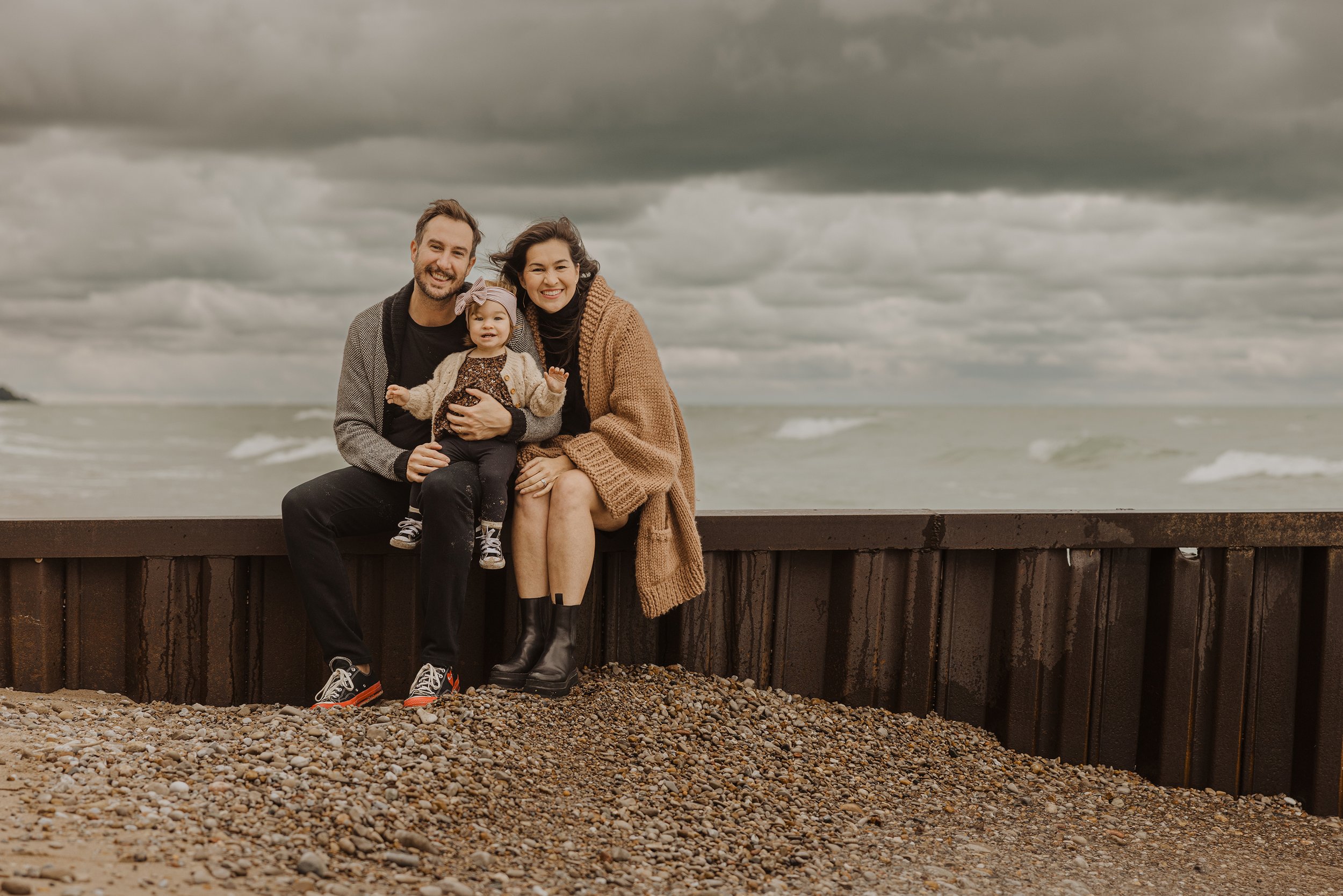 A family of three sitting on a beach with a backdrop of a cloudy sky, smiling warmly at the camera, with their young daughter in the middle.
