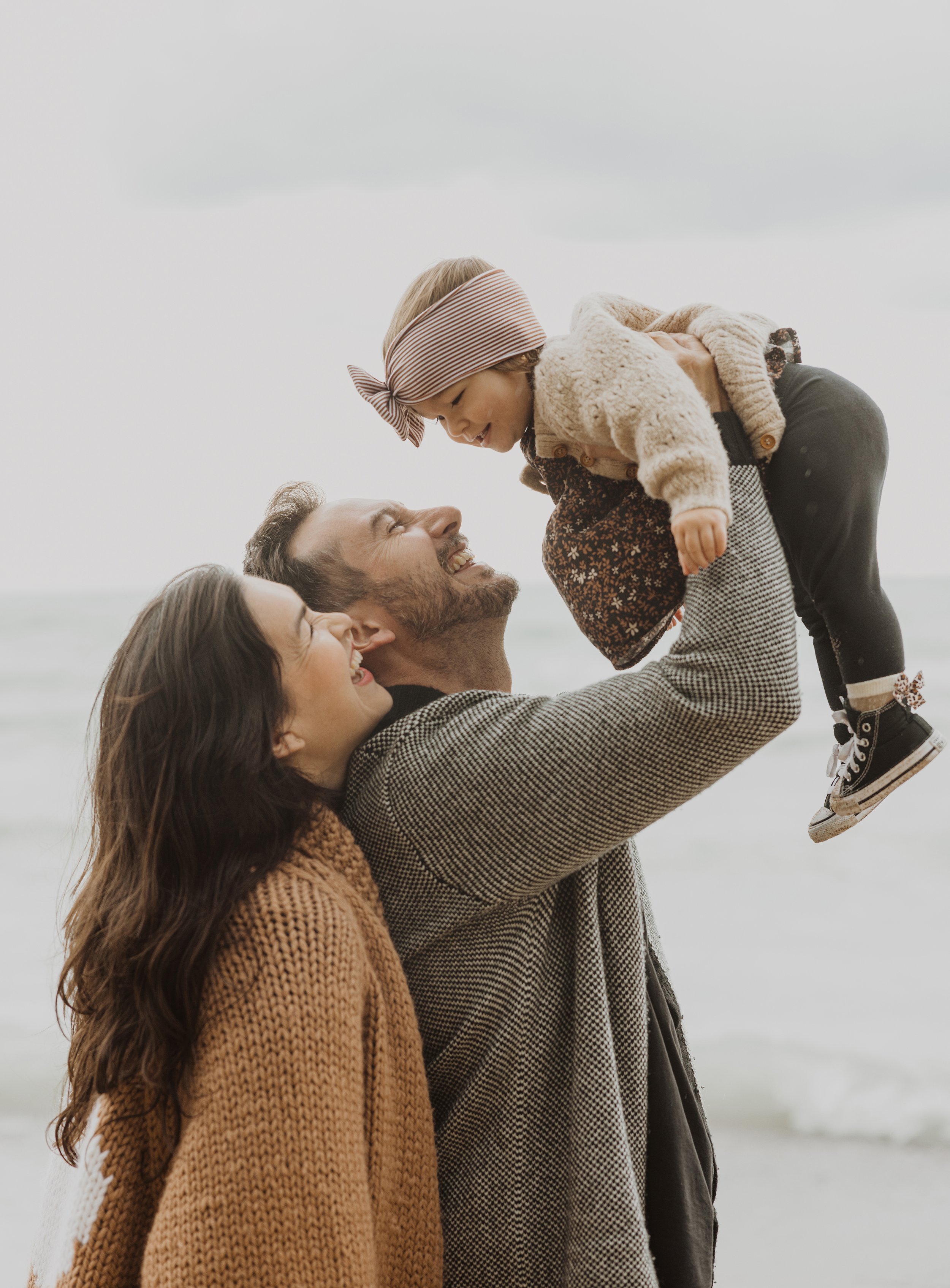A joyful moment of a father lifting his daughter playfully in the air, with the mother smiling by their side on a windy beach day.