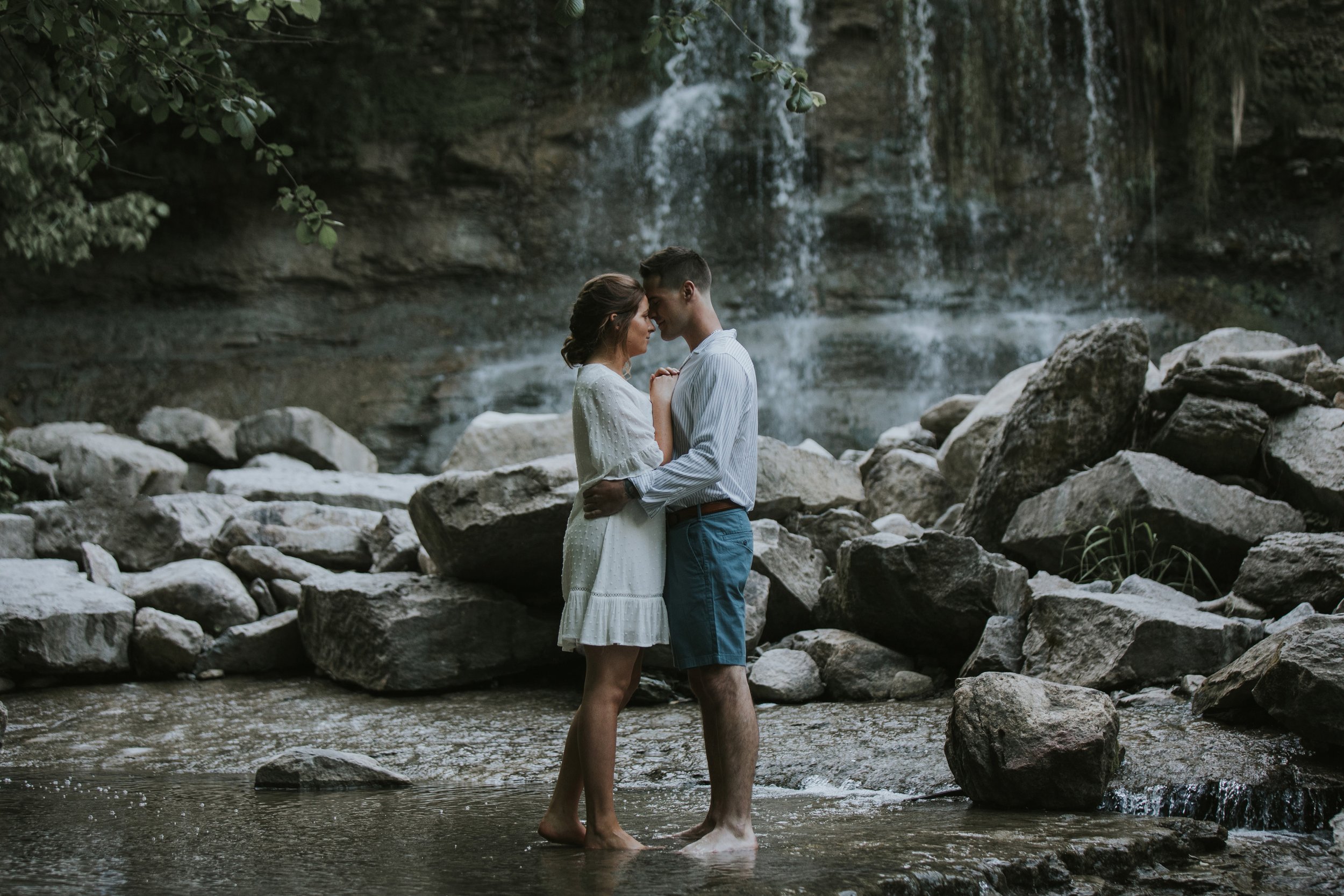 A couple shares an intimate moment by a waterfall, standing barefoot on rocks with serene expressions, surrounded by the natural beauty of cascading water and lush greenery.