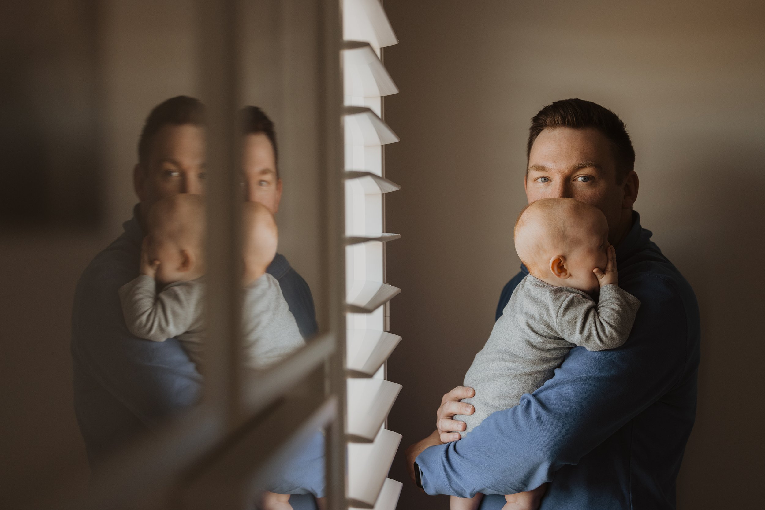 A father tenderly holding his baby by a window, with their reflection softly visible in the glass.