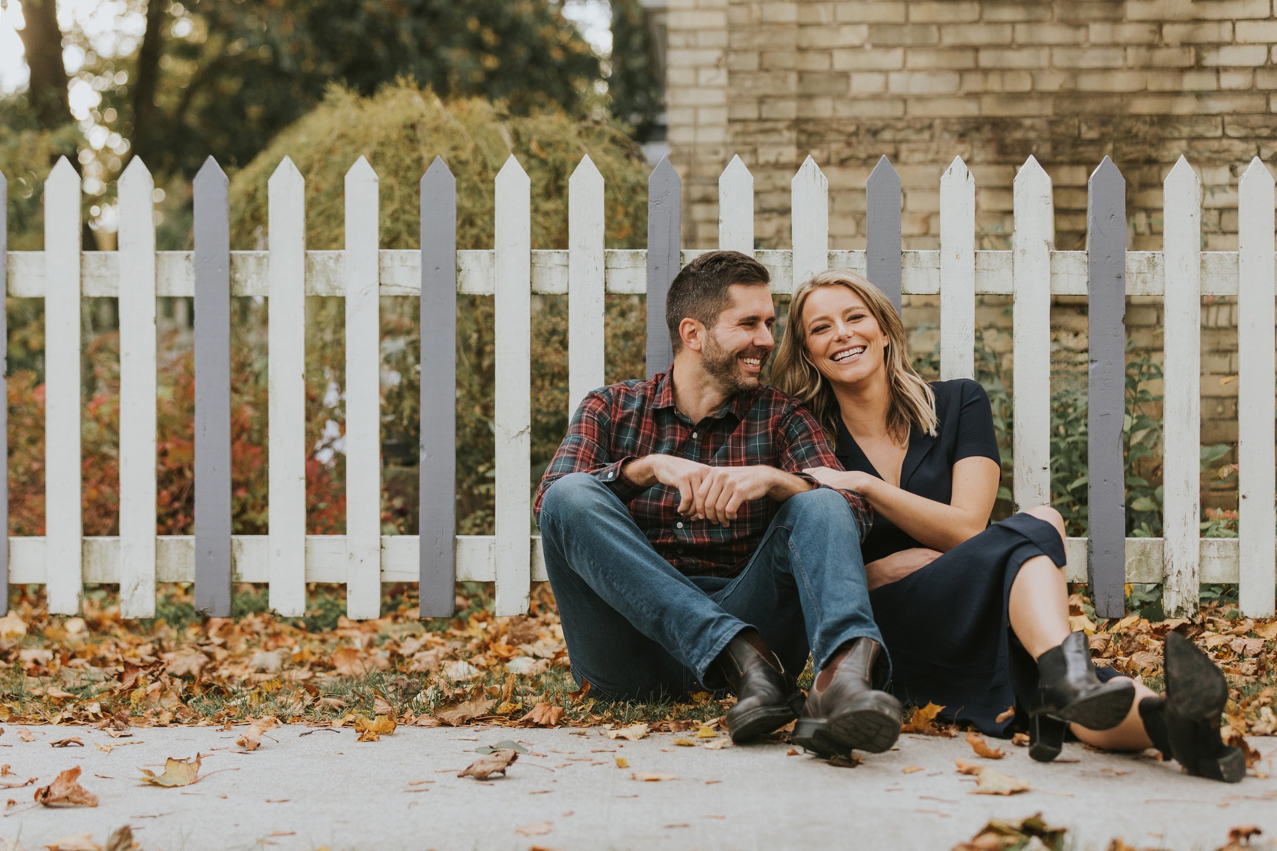 A couple sits together, laughing and leaning on each other, in front of a white picket fence surrounded by fallen autumn leaves.