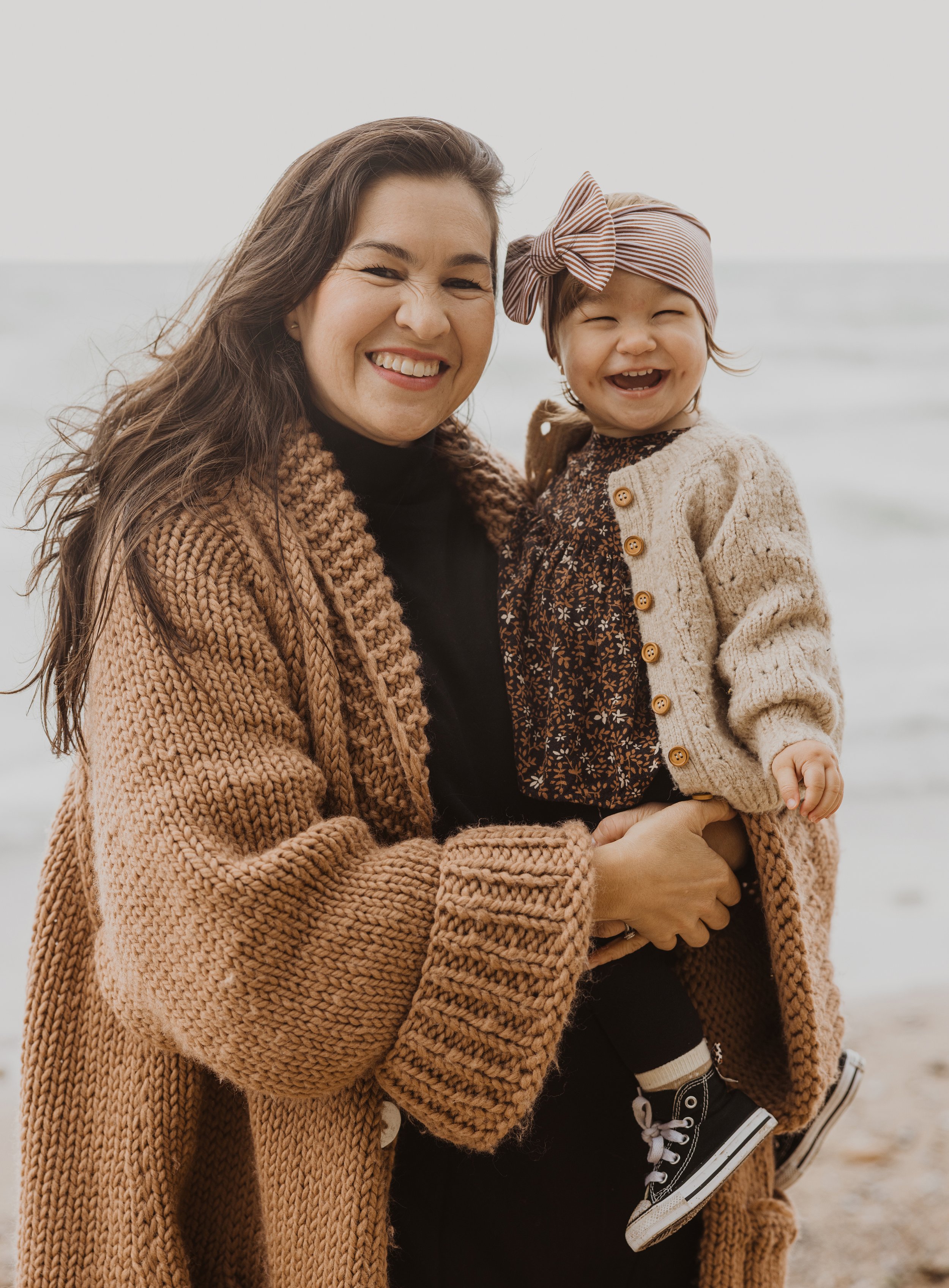 Mother and daughter sharing a joyful smile on a beach, both wrapped in cozy knit sweaters.