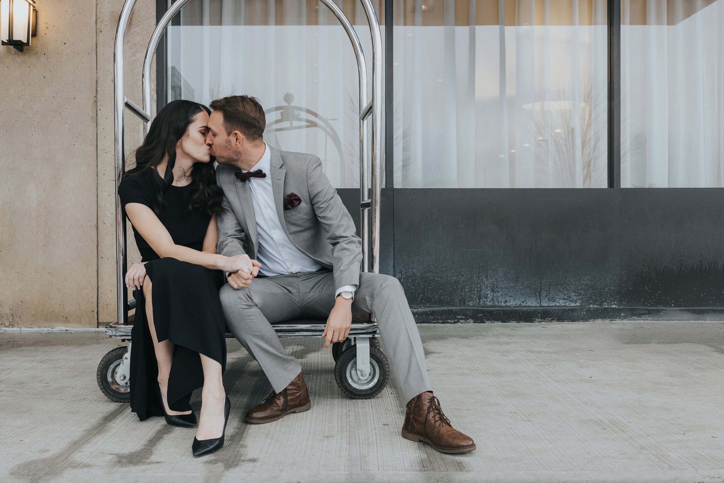 Couple kissing on a hotel sidewalk, dressed in chic, elegant attire, with the woman in a black dress and the man in a grey suit.