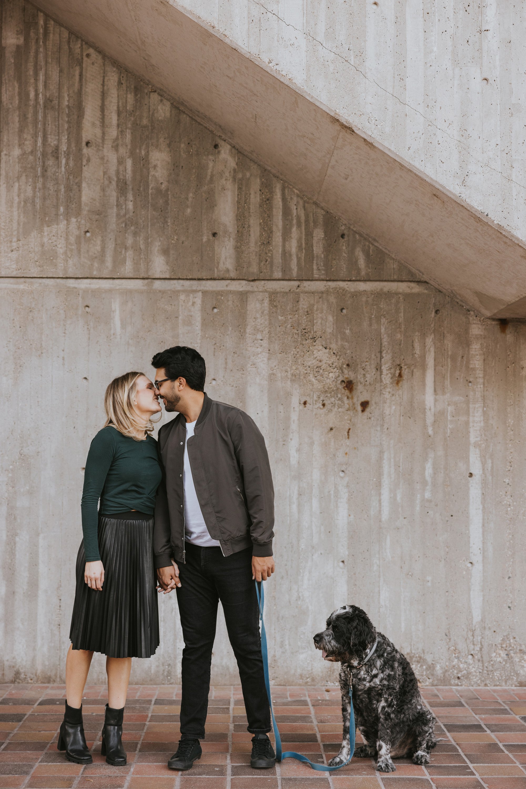 A couple with their dog shares a kiss in front of a concrete wall, combining modern and natural elements.