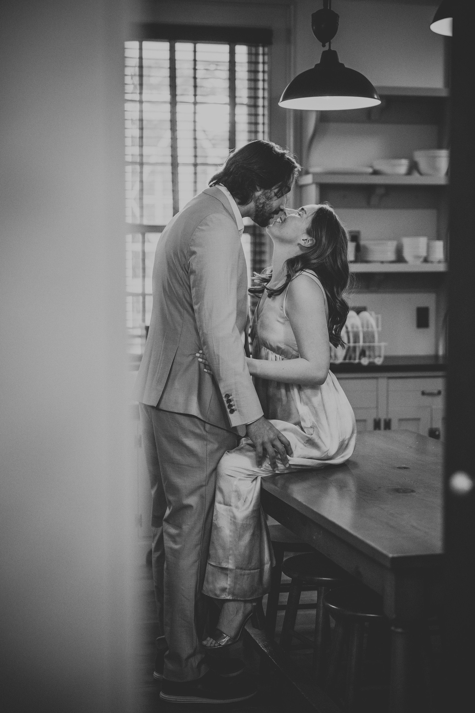 A black-and-white image of a couple sharing a romantic kiss in a cozy home kitchen.