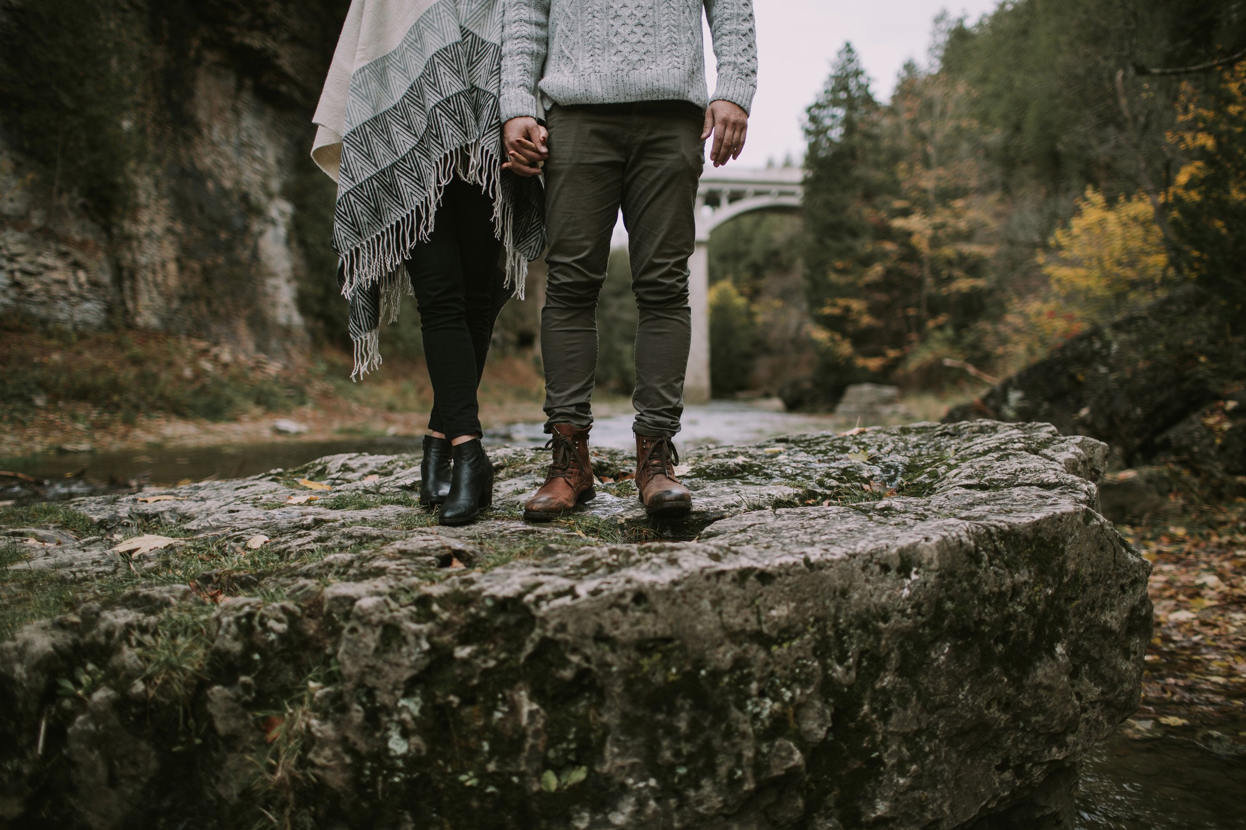 Close-up of a couple holding hands while standing on a rock near a bridge, with autumn foliage in the background.