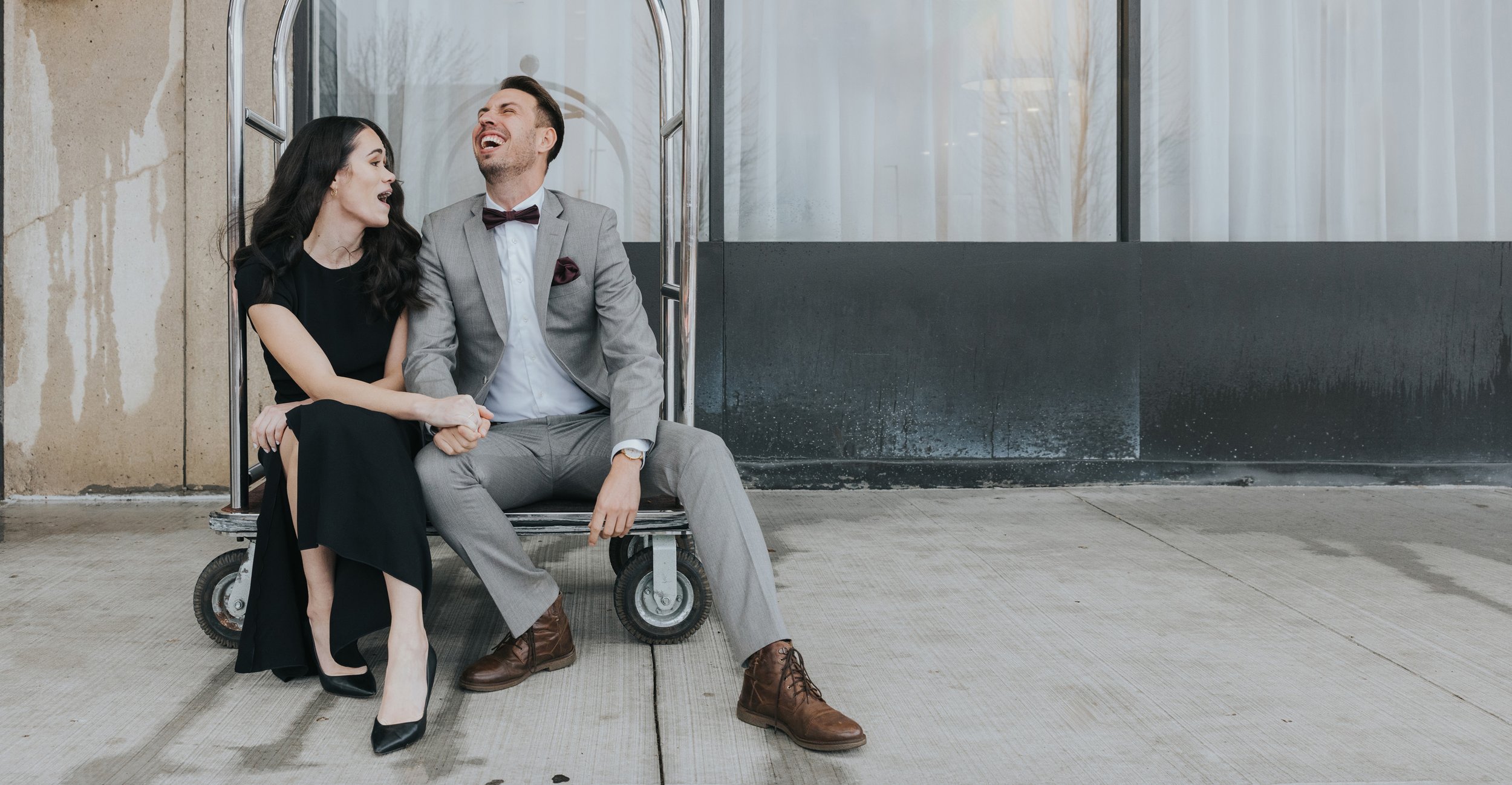 A candid shot of a couple laughing together while sitting on a hotel bell cart, enjoying a playful moment.